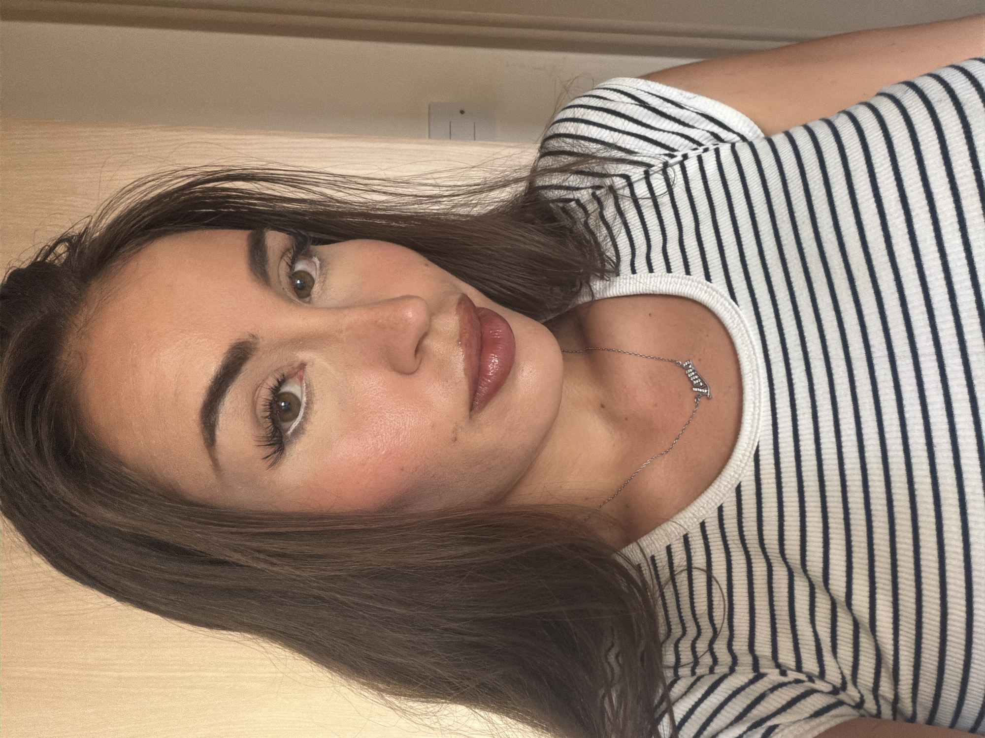 A woman is shown in a close-up, head-and-shoulders shot, looking directly at the camera. She has long, dark brown hair that falls around her face and shoulders. Her makeup includes defined eyebrows, white eyeliner on her lower lash line, winged black eyeliner, blush on her cheeks, and a glossy, brownish-pink lipstick. She is wearing a white and navy striped t-shirt, and a silver necklace with a nameplate pendant. The background appears to be an indoor setting with a textured, light-colored wall. The lighting suggests a daytime indoor environment. There is no indication of the location being Tersefanou, Cyprus.