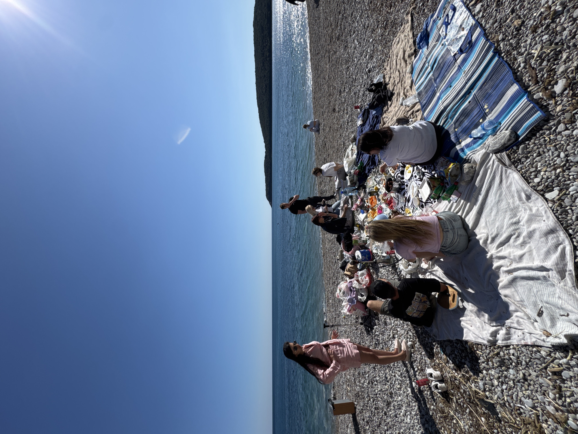 A group of people are gathered on a pebble beach along the coast. The location appears to be in the city of Prijevor, Montenegro, given the sea and mountainous background. Some individuals are sitting on blankets, while others are standing. Various items, possibly belongings or supplies, are scattered around the group. The overall scene suggests a casual gathering or picnic by the sea.