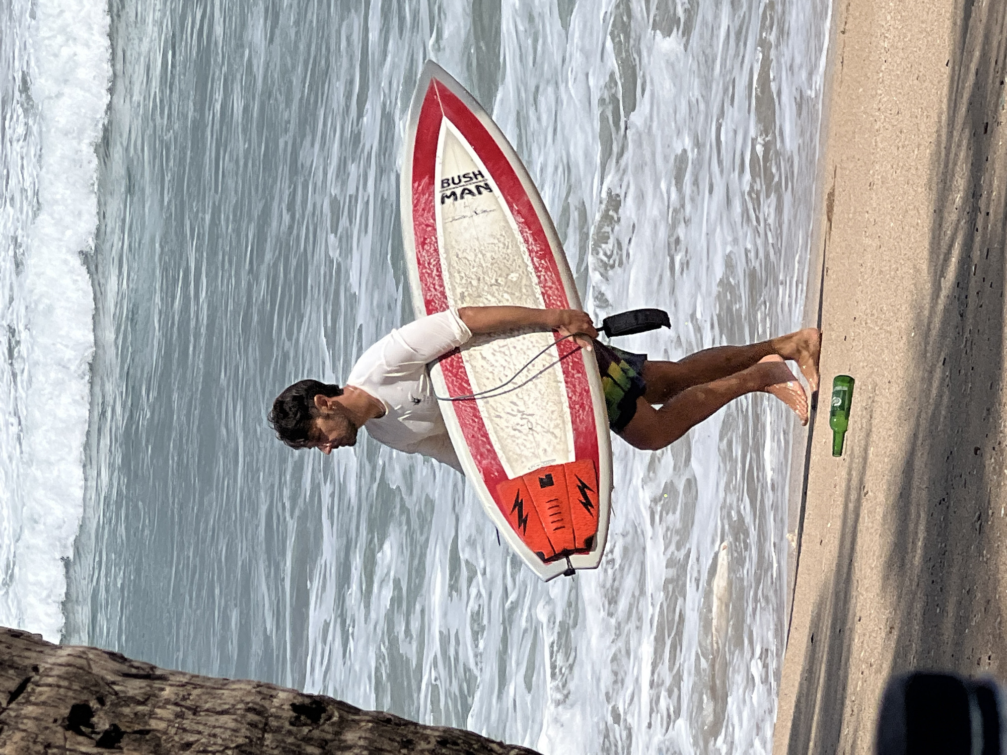 In Kuta, Indonesia, a man with dark hair and a beard, wearing a white long-sleeved shirt and multi-colored swim shorts, walks along the beach carrying a red and white surfboard. He is moving from left to right, heading towards the ocean. The surfboard has the words "BUSH MAN" printed on it. His left hand holds the leash of the surfboard. A green bottle lies on the sand in the foreground, to the left of the man's feet. The ocean is visible behind him with white waves crashing. A portion of a tree trunk is visible on the far left of the image, casting a shadow on the sand.