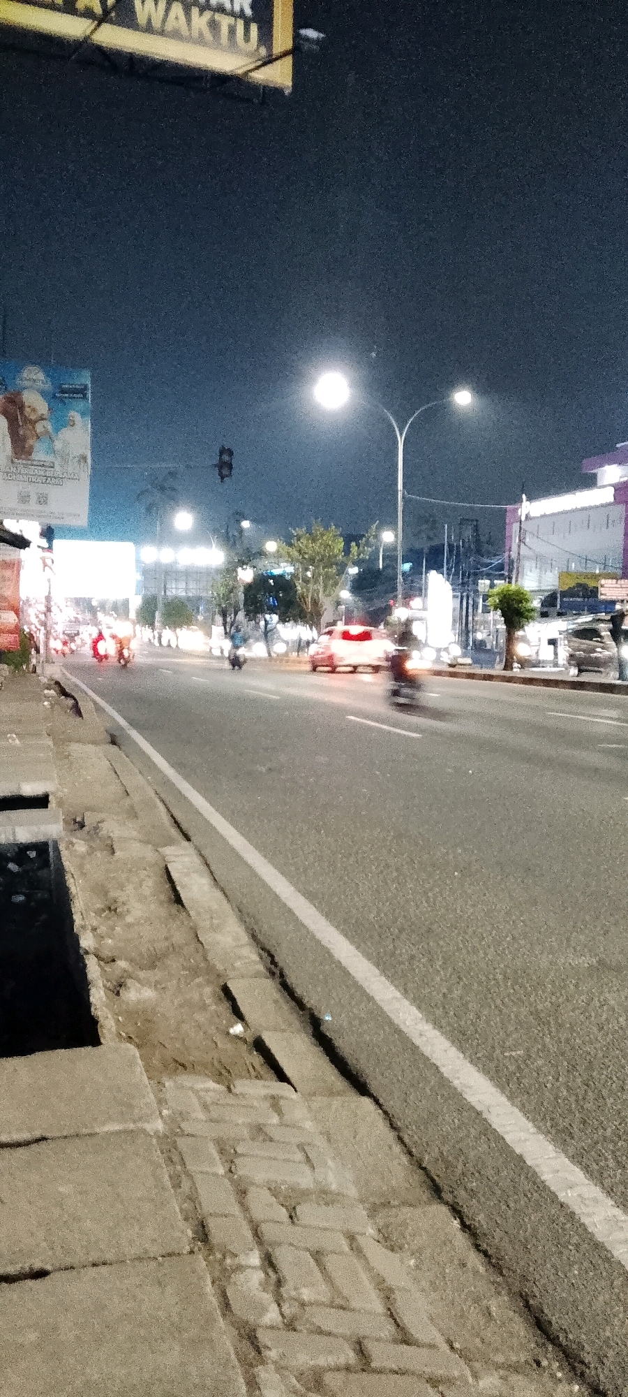 This is a night photograph of a street scene in Bekasi, Indonesia. The perspective is low, capturing the side of the road and the asphalt street.
On the left side of the image, a concrete sidewalk and drainage ditch are visible, with some damaged paving stones. Further down the street, a billboard with text in Indonesian, likely advertising a product or service, is partially visible.
The street itself is busy with traffic, primarily motorcycles and cars, indicated by the blurred lights of moving vehicles. Streetlights illuminate the scene, casting a glow on the road and the surrounding buildings. In the background, trees and modern buildings with lights can be seen. A traffic light is also visible in the distance. The overall atmosphere is that of a typical urban environment at night.