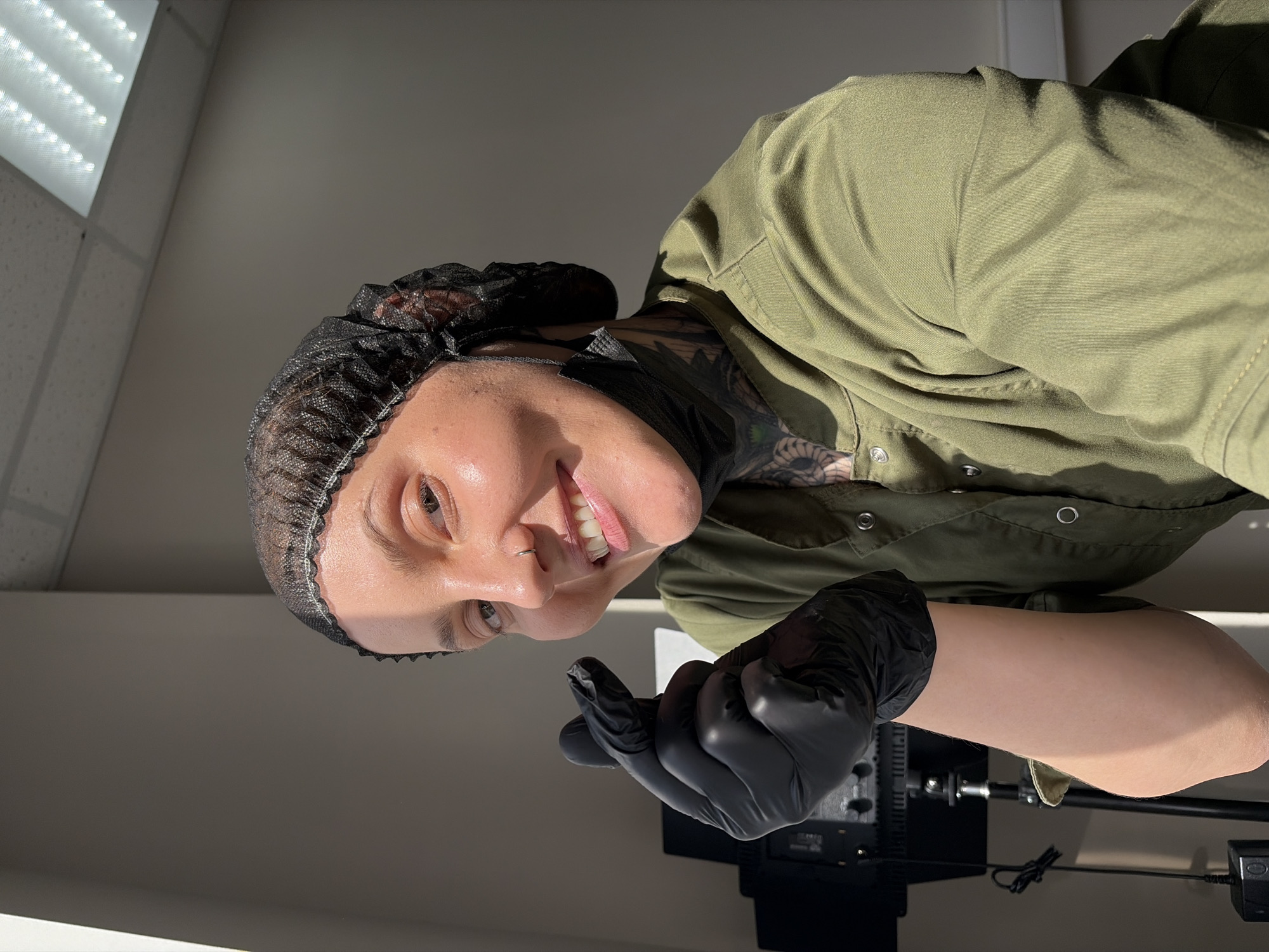 A person wearing a black hair net, black gloves, and a green scrub suit is pictured in an indoor setting. They are smiling and making a hand gesture with their right hand. A black disposable face mask is partially visible hanging from their neck. The background consists of a plain wall and a ceiling with fluorescent lighting. To the left, a black light fixture on a stand is visible. The image does not contain any information about the city of Chaikovsky, Russia.
