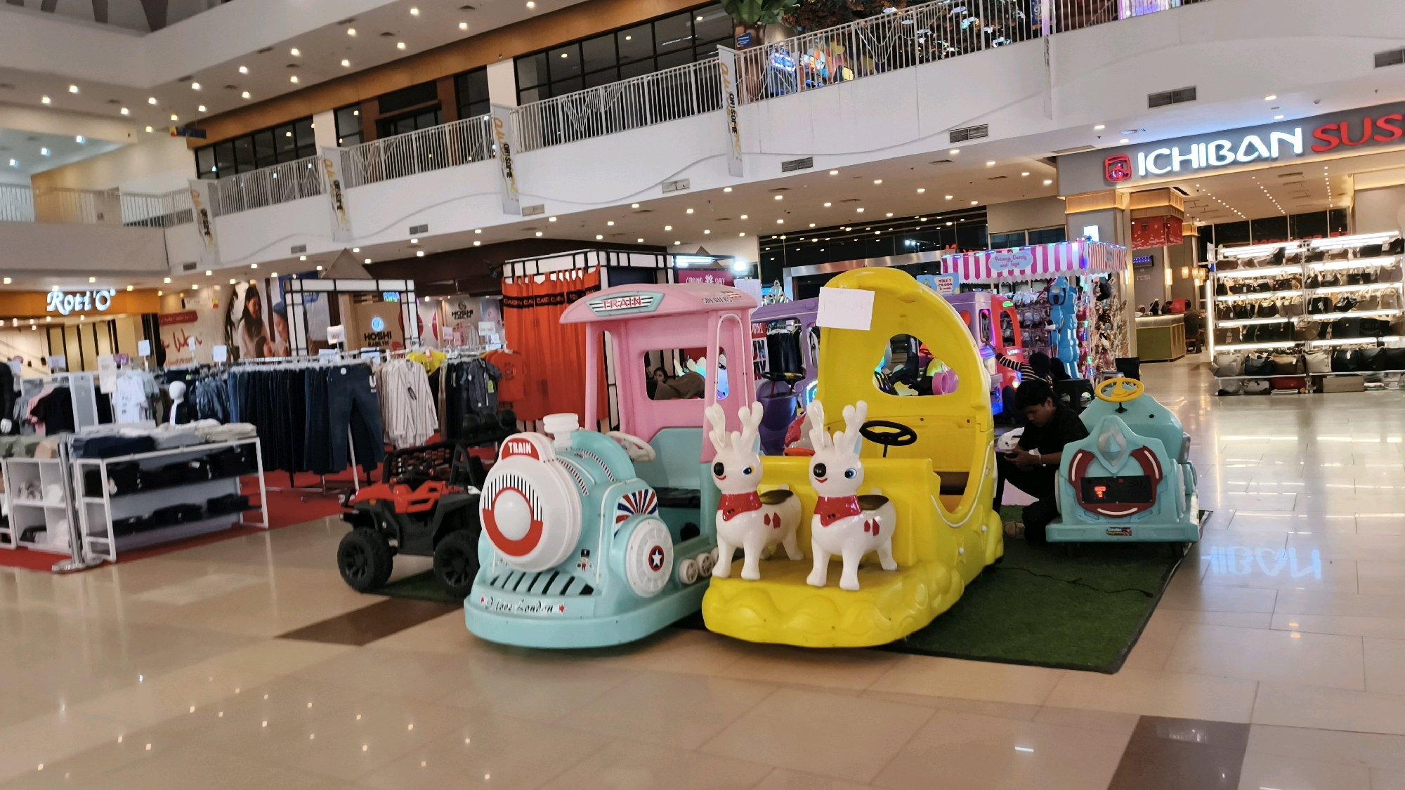 This is a wide, eye-level, interior shot of a shopping mall in Baturaja, Indonesia, featuring several children's amusement rides and store displays. In the foreground, there are three colorful, plastic amusement rides designed to resemble vehicles: a light blue train engine with "TRAIN" and "I love London" written on it, a bright yellow ride with a steering wheel and two decorative white reindeer with red scarves, and a light blue ride with a cartoon face. A young man in a black shirt is seated near the yellow and light blue rides, appearing to be interacting with his phone. Behind the rides, clothing racks filled with various garments are visible, with a store sign reading "Roti O'" in the background to the left. Further back, on the right side of the image, a store with the "ICHIBAN SUSHI" logo is prominent, displaying rows of bags on illuminated shelves. The mall's interior is well-lit with recessed lighting and features a white railing on an upper level. The floor is polished and reflects the overhead lights.