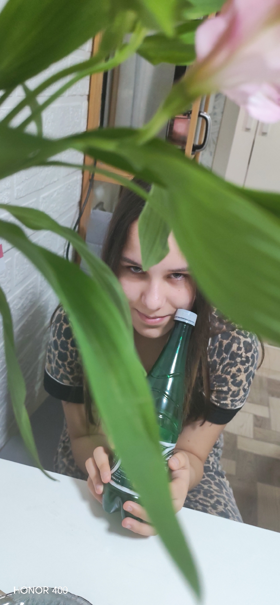 A young woman, partially obscured by green leafy plants, is holding a green water bottle. She has long brown hair and is wearing a leopard-print top. The scene appears to be indoors. A white brick wall and wooden frame are visible in the background. The location is in the city of Noviy, Russia. A white table is visible at the bottom of the frame.