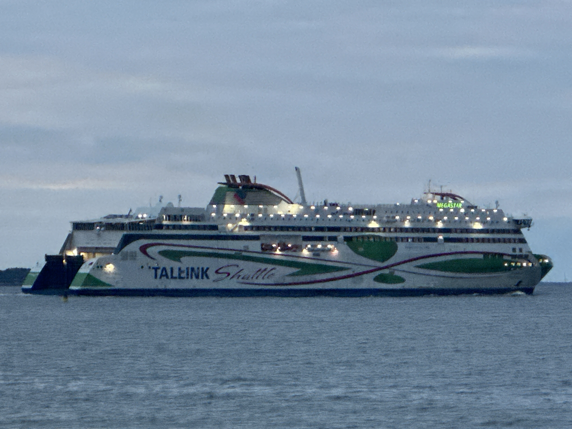 A large white and green ferry is sailing on the water. The ferry has multiple decks with many lit windows. On the side of the ferry, "TALLINK Shuttle" is written in large blue letters, and "MEGASTAR" is written in green letters on the top deck. The ferry has a white hull with green and red stripes. The water is calm and appears to be a dark blue or gray color. The sky is overcast and a pale blue or gray, suggesting it is either early morning or late evening. There are no people visible on the ferry or in the water. The scene appears to be a serene and quiet moment at sea, possibly near a port in Tallinn, Estonia, as indicated by the ferry's company name.