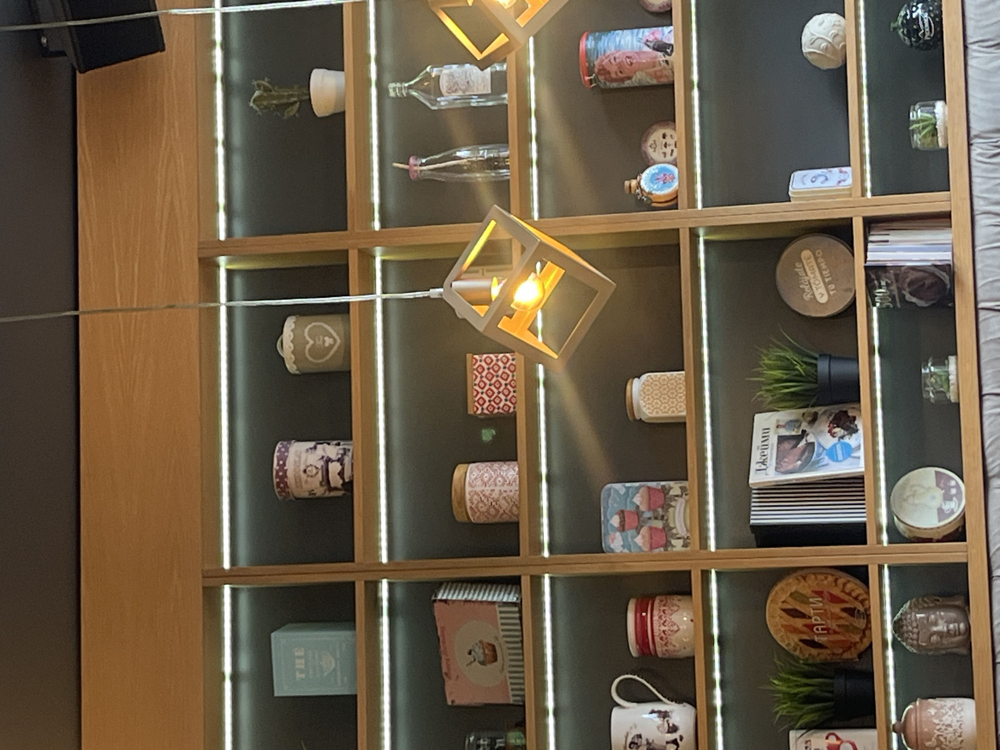 The image presents a wall-mounted shelving unit, constructed of light-colored wood with dark grey backing in each compartment, displaying various decorative objects. The unit is illuminated with vertical light strips between each compartment.
The shelving contains a variety of items including: plants in pots, decorative jars and bottles, mugs, books, decorative boxes, and small sculptures. Some objects appear to be vintage or antique.
A modern, cube-shaped pendant light fixture is suspended in front of the shelving, with the light bulb visible. The light hangs in the foreground of the shelving.
The image does not explicitly indicate a location in Veliko Tarnovo, Bulgaria. However, the composition suggests an interior design context, possibly within a shop, cafe, or private residence.
