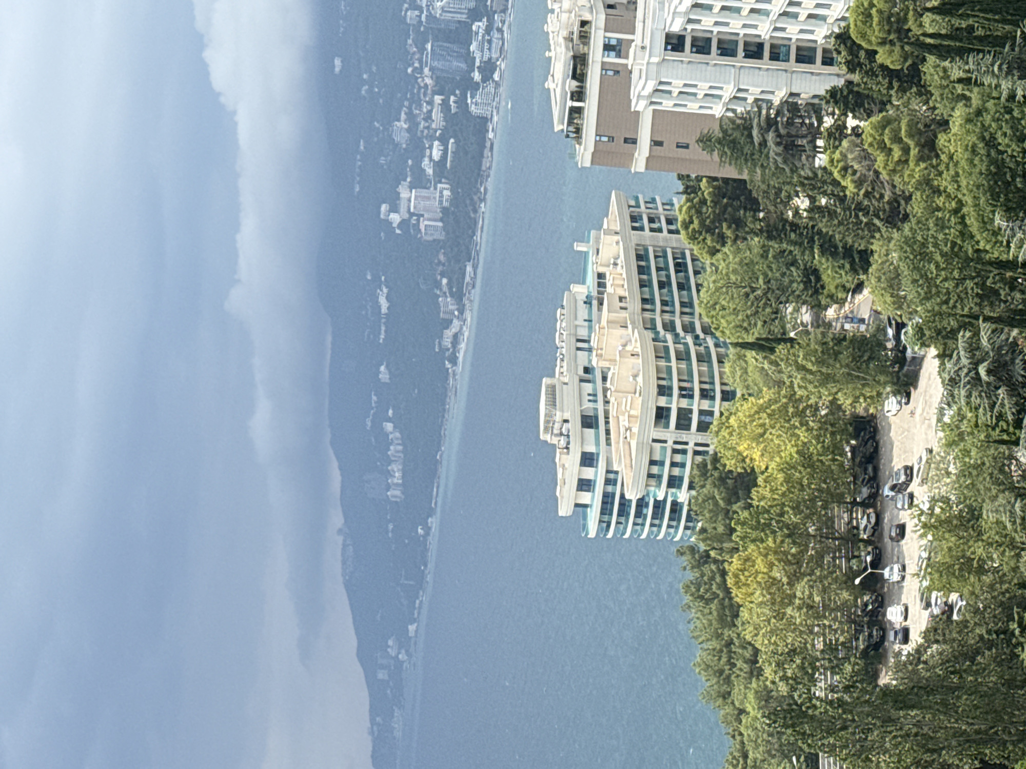 An elevated, wide shot captures a coastal cityscape under a cloudy sky. In the foreground, lush green trees dominate, with a parking lot filled with approximately 20 cars visible amidst the foliage. Two modern, multi-story buildings with rounded balconies and light-colored facades are situated on the hillside, overlooking the sea. Further in the distance, the turquoise ocean stretches out to meet a hazy, mountainous coastline dotted with numerous buildings, suggesting a dense urban area. The overall scene depicts a resort town with residential and possibly hotel structures overlooking the sea, framed by abundant vegetation.