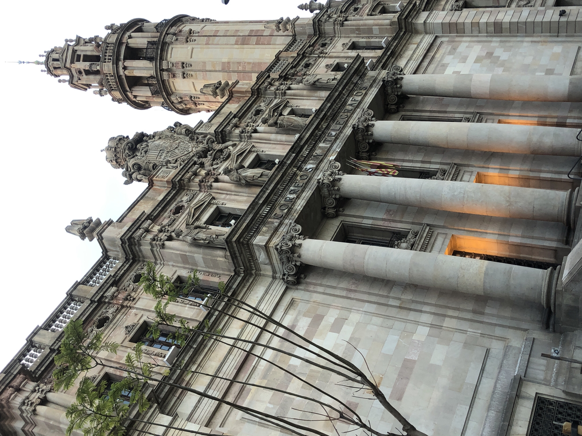 The image shows the facade of a large, ornate building constructed of stone. The building has classical architectural elements, including large columns, sculptures, and intricate carvings. There are multiple windows on the facade, some of which are illuminated from within, casting a warm glow. A tower with a spire rises from the right side of the building. In front of the building, on the left side of the image, are several thin, bare tree branches. The sky above is bright and overcast. The architectural inscription "CORREOS Y TELEGRAFOS" is visible above the columns. Two flags, one with yellow and red stripes, hang from a window.