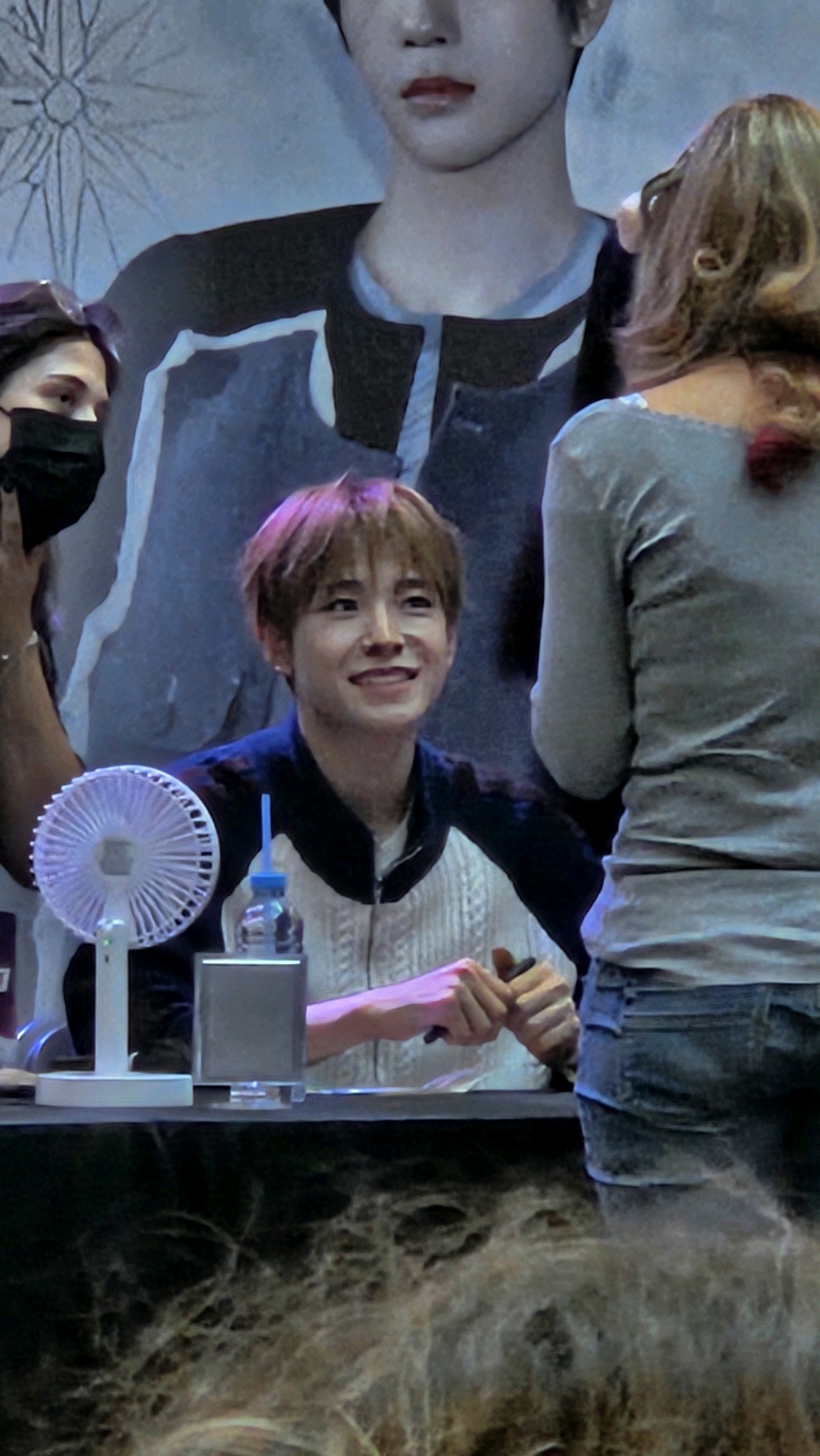 The image shows a close-up of a young man with short, light brown hair, smiling at the camera. He is wearing a black and white jacket with a white shirt underneath. To his left, a woman with dark hair is wearing a black face mask. To his right, another person is seen from the back, wearing a light grey shirt and denim shorts. In front of the young man are a small white fan and a clear bottle with a blue straw. The background appears to be a large poster or mural of a person's torso and head. The scene is likely indoors, possibly at an event or fan meeting. There is no indication of the city of Barcelona, Spain.
