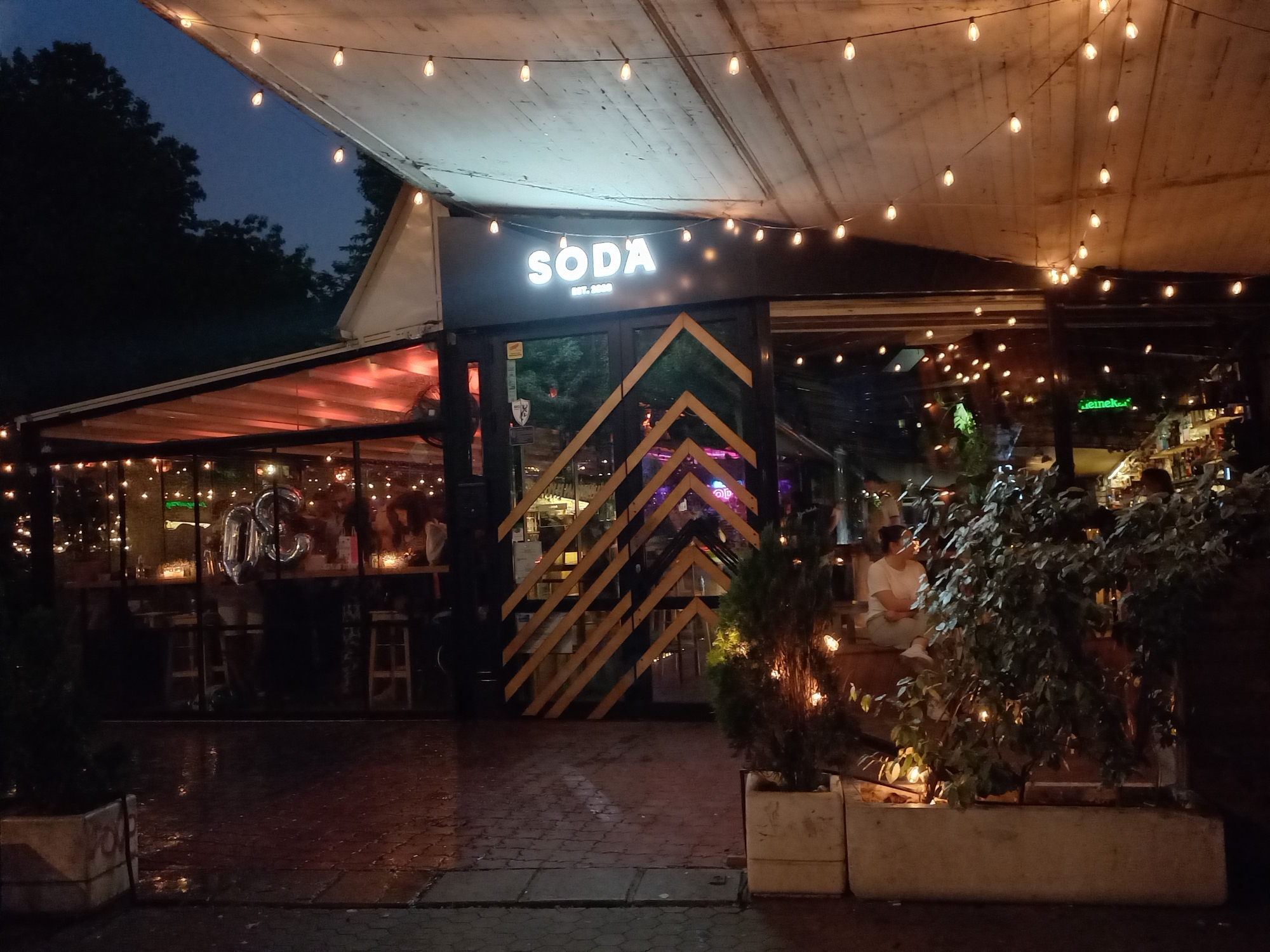 The image displays the illuminated exterior of a bar or restaurant named "SODA Est. 2020" at night, set against a dark blue twilight sky with silhouetted trees in the background. The scene depicts a lively establishment, likely located in Sofia, Bulgaria, characterized by warm ambient lighting.
A white, possibly canvas, canopy extends over the front of the building and an outdoor seating area, featuring numerous string lights with bare bulbs that cast a warm glow on the wooden ceiling panels. The main sign, "SODA Est. 2020," is prominently displayed in bright white letters above the entrance. The building itself is enclosed primarily by large glass panels, allowing visibility into the interior where more string lights, patrons, and a "Heineken" logo are discernible. Inside, near a counter, a large silver balloon resembling "30" or "OC" is visible.
The main entrance features a distinctive pattern of diagonal wooden slats in alternating black and light brown tones. In front of the building, the ground is paved with dark bricks, appearing somewhat damp and reflecting the scattered lights. Planters containing small trees and shrubs, also adorned with small lights, flank the entrance and seating areas, adding to the decorative effect. To the right, outside the main entrance, a person in a white top and light trousers is seated on a ledge amidst the greenery, looking towards the right. The overall atmosphere is inviting and social.