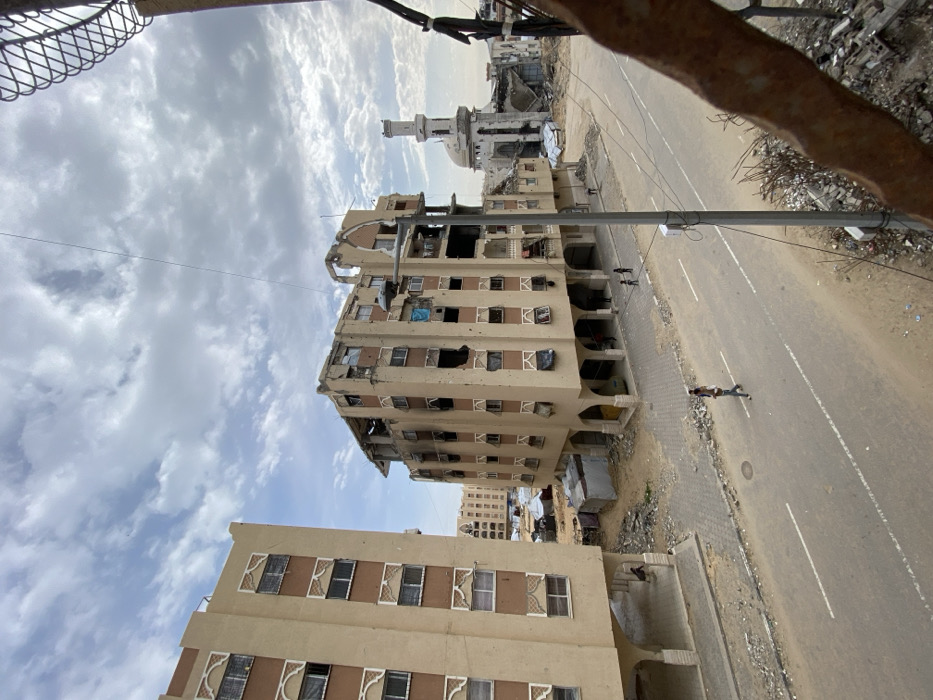An elevated view depicts a heavily damaged urban landscape in Khan Yunis, Palestinian Territory, under a partially cloudy sky. In the foreground, a rusty, gnarled metal bar occupies the extreme right, with a pile of concrete rubble directly below it.
An asphalt road runs through the mid-ground, featuring two dashed white lane markers. An adult, carrying a child on their shoulders, walks leftward on this road. Further down, two smaller figures are visible on the left sidewalk.
To the left of the road, a multi-story, beige residential building with brown trim and decorative window elements stands. It shows some missing or covered windows but appears largely structurally intact, with arched open supports on its ground floor.
In the center, another multi-story, beige building exhibits extensive damage. Its facade is riddled with numerous impact holes, consistent with bullet or shrapnel strikes. Significant portions of its upper floors and roofline are destroyed, and many windows are shattered or missing, revealing damaged interiors; some window openings are covered by blue tarps. The ground floor also features arched supports.
Between these two buildings, an open, sandy area contains several small, makeshift tent-like structures. Further in the background, a mosque with a distinct white dome and a partially damaged, slender minaret is visible amidst substantial surrounding structural wreckage. A grey street light pole with a modern fixture stands on the right side of the road, supporting overhead wires.