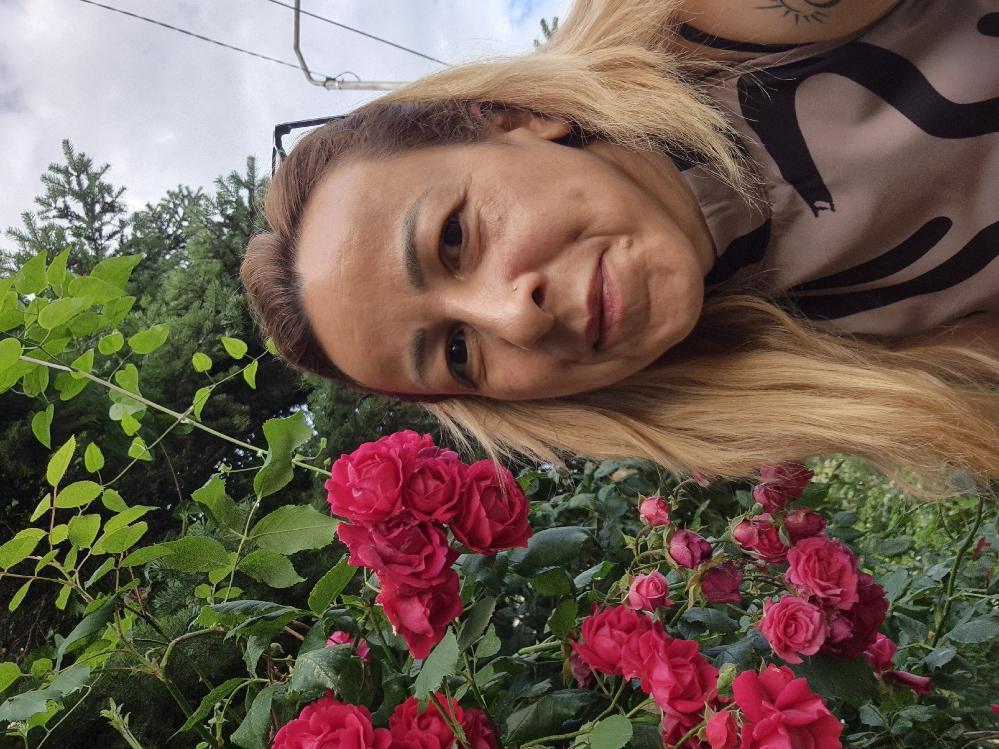 The image captures an outdoor portrait of a woman standing to the right of a vibrant rose bush. The woman has shoulder-length, blonde to light brown hair, parted slightly to her right, with strands falling over her shoulders. She has a small silver stud piercing on the left side of her nose and a small raised mole below her lower lip. She is looking directly at the camera with a subtle smile. Her left shoulder and upper arm are visible, revealing a tattoo of a stylized sun. She is wearing a sleeveless, high-necked top that is light brown or beige with a bold, abstract dark brown or black pattern, including a design resembling a snake's head on her upper chest. Glasses are perched on top of her head, partially visible above her forehead.
To the woman's left, a dense rose bush with numerous dark pink to red blooms and green foliage occupies the left and bottom-center portions of the image. Many roses are in full bloom, while others are tightly closed buds, and some show signs of wilting. Behind the woman and the rose bush, a mix of dark green foliage from trees and bushes is visible. The upper right portion of the background shows an overcast, cloudy sky. A light-colored metal pole or structure with attached wires is visible against the sky on the far right edge of the frame. The scene takes place outdoors in the city of Unknown, Unknown.