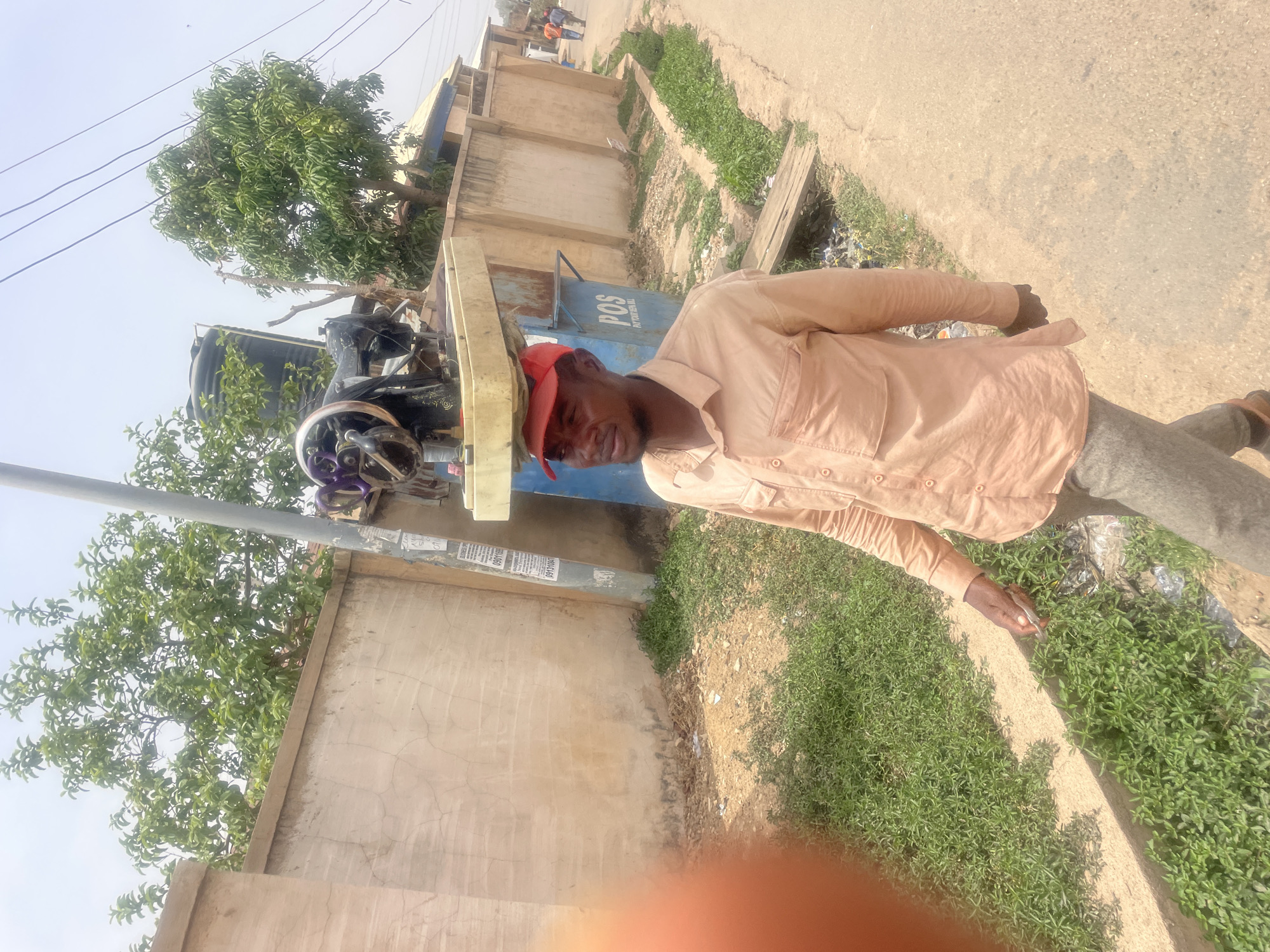 The image, initially rotated 90 degrees clockwise, captures a bright, sunny outdoor scene, likely in a semi-urban area of Jos, Nigeria.
The central subject is a dark-skinned man, wearing a light peach/tan long-sleeved collared shirt, grey trousers, and a red baseball cap. He is standing, looking directly at the camera with a neutral expression.
To his left (when the image is viewed upright) is a vintage black sewing machine, mounted on a light-colored wooden stand. Behind the sewing machine is a blue structure or kiosk with the clear text "POS" and "PAYMENT HALL" visible on its side. Further behind these objects, a large, dense green tree and a black water tank can be seen. A utility pole is situated near the sewing machine, adorned with several stapled or taped paper notices, some showing partial numbers like "080345...". Overhead, thin power lines stretch across a clear, bright sky.
The man is positioned at the edge of a dirt and concrete path or roadside. To his right, the ground transitions from a bare earth patch to an area covered with short, vibrant green grass. In the distance down the street, two smaller figures are visible, seemingly walking. The scene is illuminated by harsh sunlight, suggesting midday. A blurry orange/red object, possibly a finger, partially obstructs the bottom right corner of the frame.
