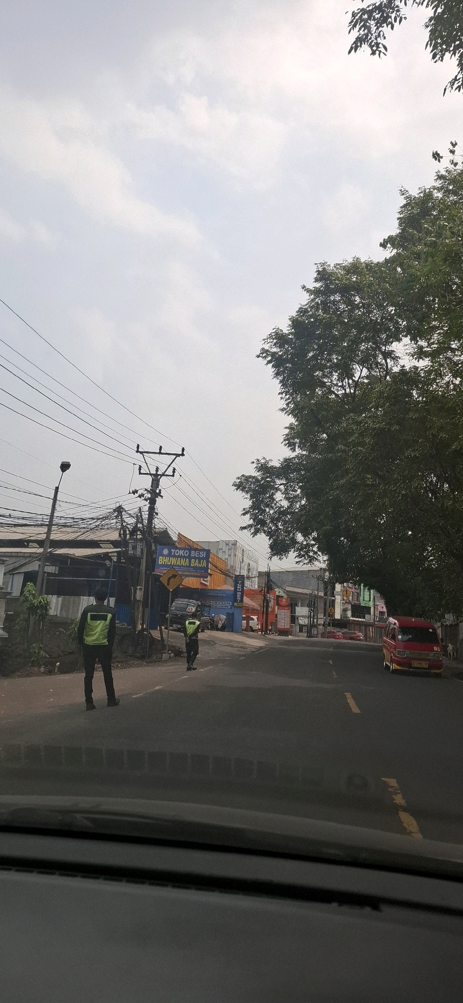 The image captures a street scene in Sirnagalih, Indonesia, under an overcast sky. On the left side of the road, two individuals wearing high-visibility vests and dark pants stand on the sidewalk. The one closer to the camera is facing away, while the other is turned slightly towards the road. Behind them, a building with a weathered facade is visible, along with a utility pole supporting numerous electrical wires. A sign for "TOKO BESI BHUWANA BAJA" (Iron Shop Bhuwana Steel) is mounted on a pole.
The road itself is paved, with a yellow line marking the center. A dark-colored car is parked on the left side of the road, and further down the street, a red van with yellow trim is parked on the right. Various buildings line the street, some with commercial signage. A large tree with dense foliage is present on the right side of the frame, its branches extending over the road. The lower portion of the image shows the interior of a vehicle, likely the vantage point from which the photo was taken, with a blurred view of the dashboard and windshield.