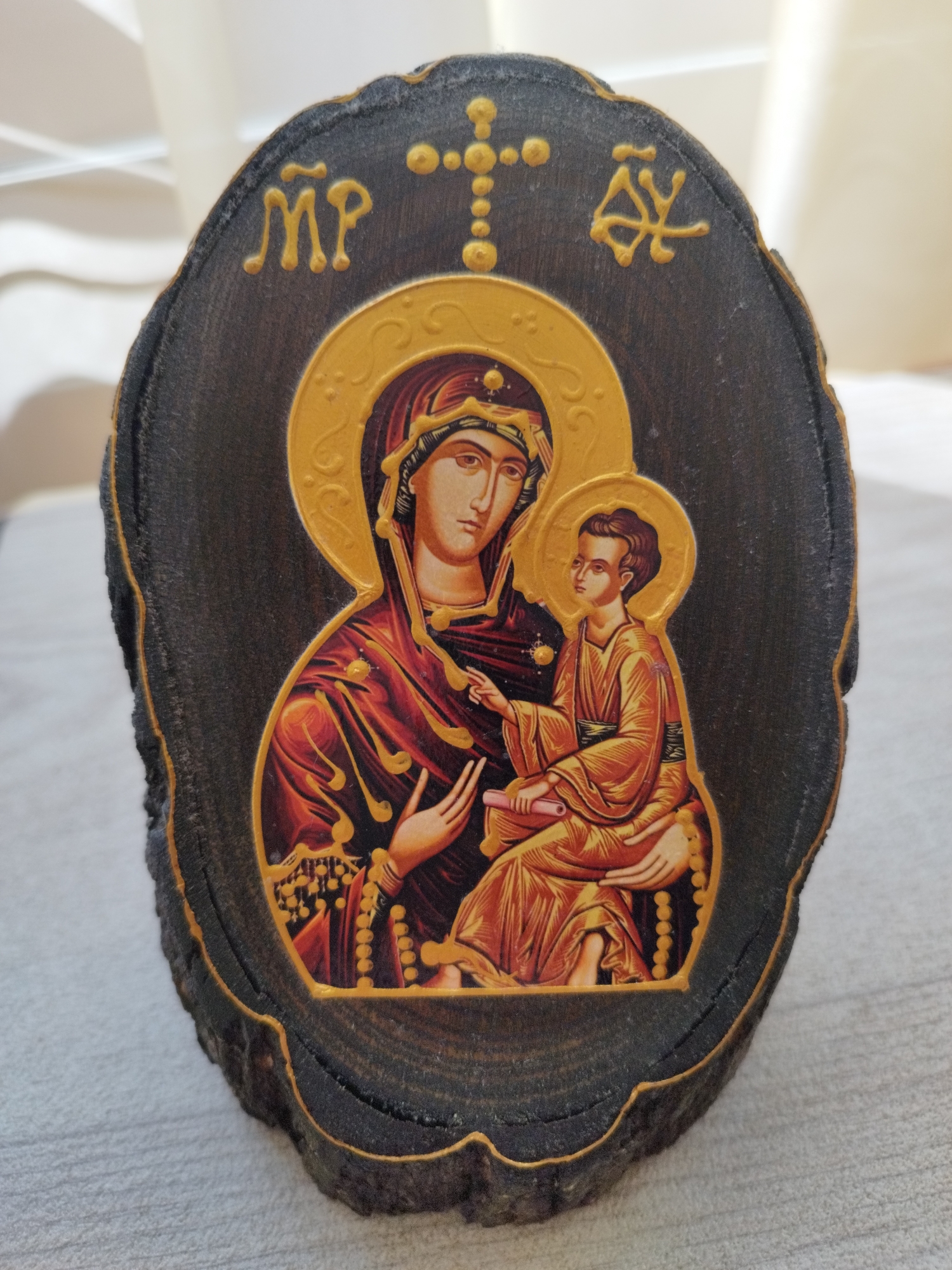 A wooden icon of the Virgin Mary and Jesus Christ is displayed on a neutral surface. The icon is an oval-shaped wood slice with the image of the Theotokos (Mother of God) holding the infant Jesus affixed to the front. Above the image, gilded characters of "MP" and "ΘΥ" are visible, as well as a cross. The style of the image is Byzantine-inspired. The bark edge of the wood slice is retained, giving the icon a rustic appearance. The background suggests an interior setting with a partially visible, light-colored fabric or surface behind the icon. It’s not possible to provide the specific location in Sofia, Bulgaria from the information provided.