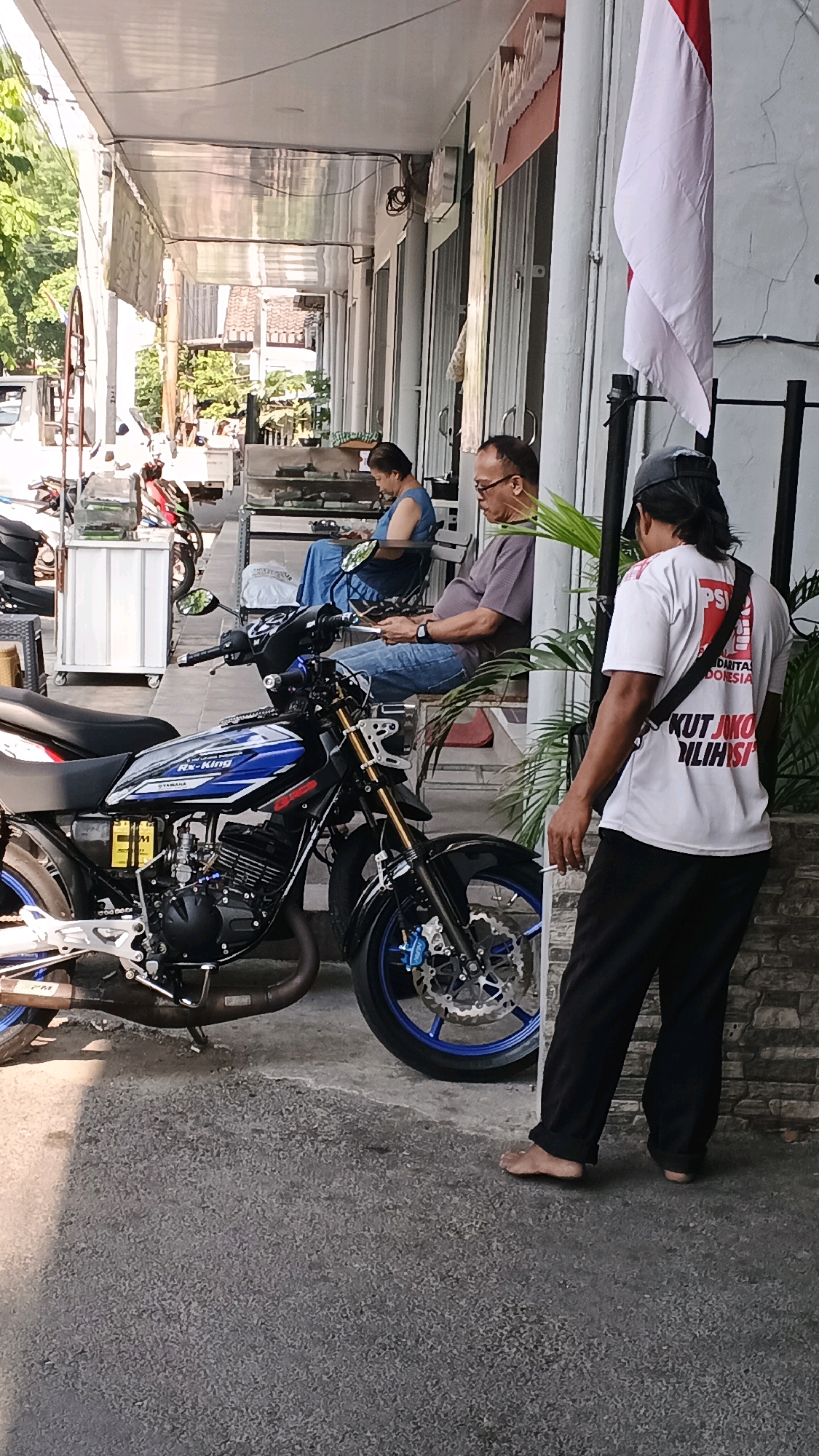 The image depicts a street scene in Semarang, Indonesia, likely outside a shop or residential building. In the foreground, a blue and black motorcycle is parked on the asphalt. The motorcycle features visible branding, including "RX-King" and "YAMAHA." To the right of the motorcycle, a man wearing a white t-shirt with Indonesian text and black pants stands barefoot, facing away from the camera. He appears to be smoking a cigarette. In the background, two individuals are seated on a bench. One, a woman in a blue garment, is looking down at her hands. The other, a man wearing glasses and a grey t-shirt, is looking at a phone. Behind them, a red and white Indonesian flag hangs from a pole. The environment includes a covered walkway, a wall with stone-like tiling, and various shopfront elements. Green foliage is visible to the right.
