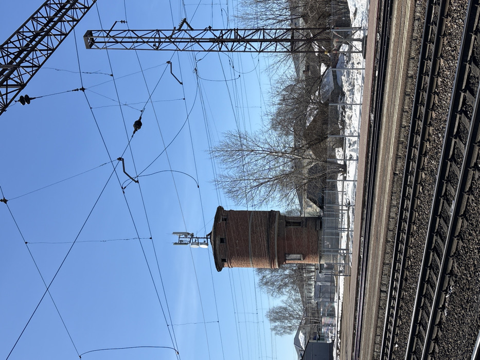 The image depicts a railway environment in Kropachyovo, Russia, under a clear blue sky. In the foreground, multiple sets of parallel railway tracks with dark gravel ballast are visible, alongside a reddish-brown concrete railway platform. A gray metal fence separates the immediate railway area from the midground.
The central subject is a cylindrical, two-story brick water tower, predominantly red-brown. Its upper section features a metal superstructure supporting multiple cellular telecommunication antennas, including panel antennas and several circular dish antennas. Windows are visible in the tower's facade. To the right of the tower, a large lattice steel pylon, part of the railway's overhead electrification system, stands tall, supporting a complex network of overhead power lines and catenary wires with associated insulators.
Bare deciduous trees, indicating a late winter or early spring season, are scattered across the midground. Patches of white snow are visible on the ground, particularly behind the fence. In the background, beyond the trees, several residential or utility buildings are partially visible under the clear blue sky. No individuals, actions, or interactions are present in the scene.