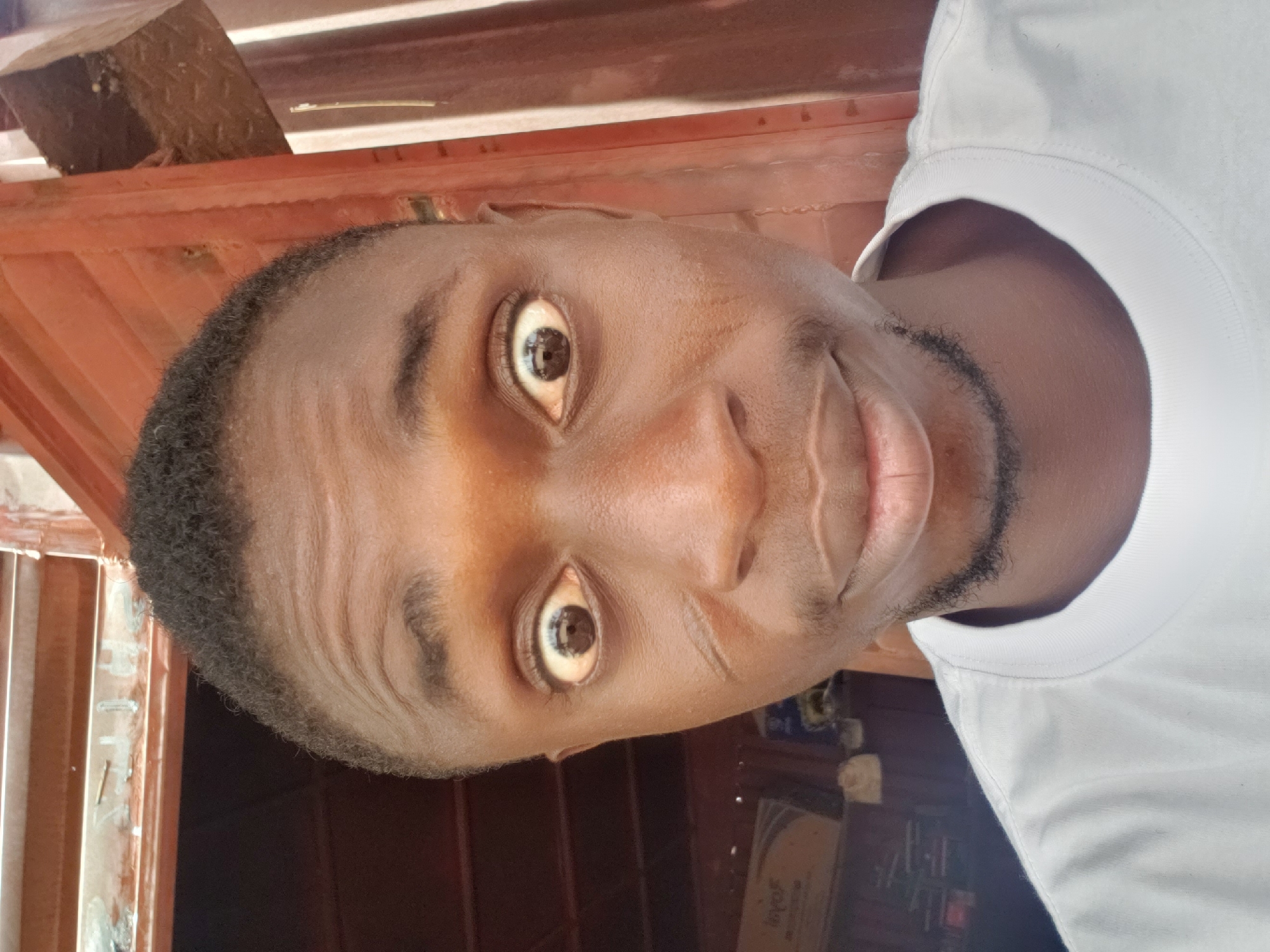 The media file displays a close-up, head-and-shoulders portrait of a young Black man, likely a selfie given the angle. He has dark skin, short black hair, and a short, dark beard outlining his jawline and chin. He is wearing a plain white round-neck t-shirt. His eyes are wide open, appearing alert or surprised, and look directly into the camera, featuring dark irises with reflections. There is a subtle hint of a smile or smirk on his lips.
The background is somewhat out of focus. Above and behind him, a reddish-brown structure is visible, possibly corrugated metal sheets or painted wooden panels, suggesting an indoor or shaded outdoor setting. To the right of his head and shoulder, the background is darker, with indistinct shapes that could be furniture or other objects in a room. The lighting indicates daylight, casting a slight shine on his forehead and nose. The image captures a direct and casual moment, conveying a sense of immediacy. The picture was taken in Adankolo, Nigeria.