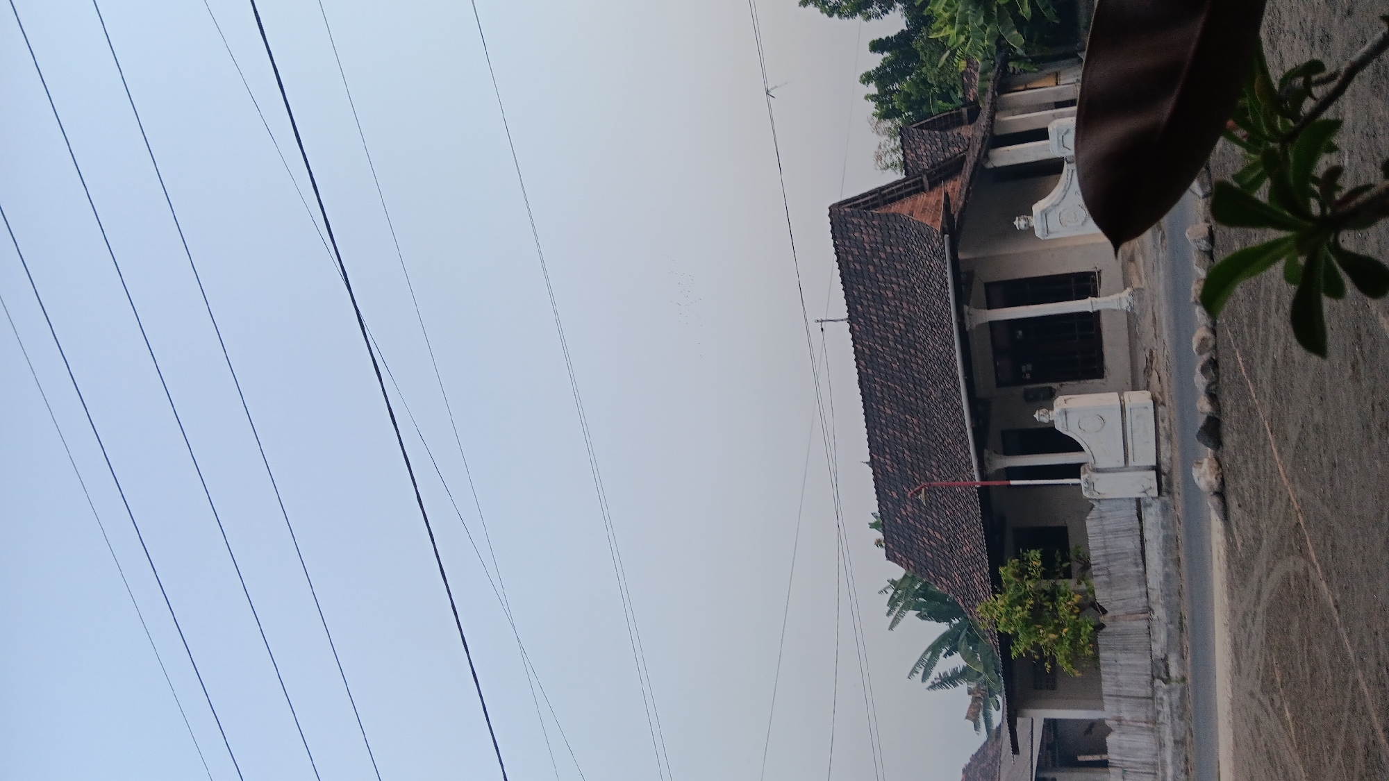 The image depicts a residential scene in the city of Sonobekel, Indonesia. The central focus is a traditional Indonesian house with a brown tiled roof and a white, columned porch. In front of the house, a white fence separates it from a road. The yard in the foreground is covered with sand and features some rocks and sparse vegetation. Overhead, several power lines traverse the sky, which is a hazy, pale blue. A flock of birds is visible in the distance, high in the sky. The overall impression is of a quiet, rural or suburban setting.