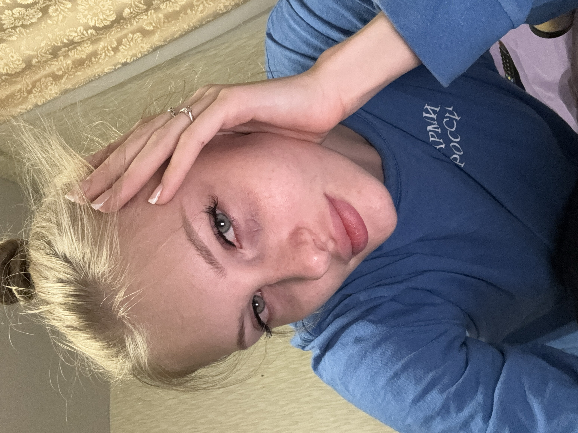 A young woman with blonde hair in a bun is looking at the camera, with her left hand resting on her forehead and her right hand on her cheek. She is wearing a blue sweatshirt with white Cyrillic lettering on the left side of the chest. The background consists of a yellow patterned curtain and a beige wall. The image appears to be taken indoors, likely in a residential setting in Kondratovo, Russia.