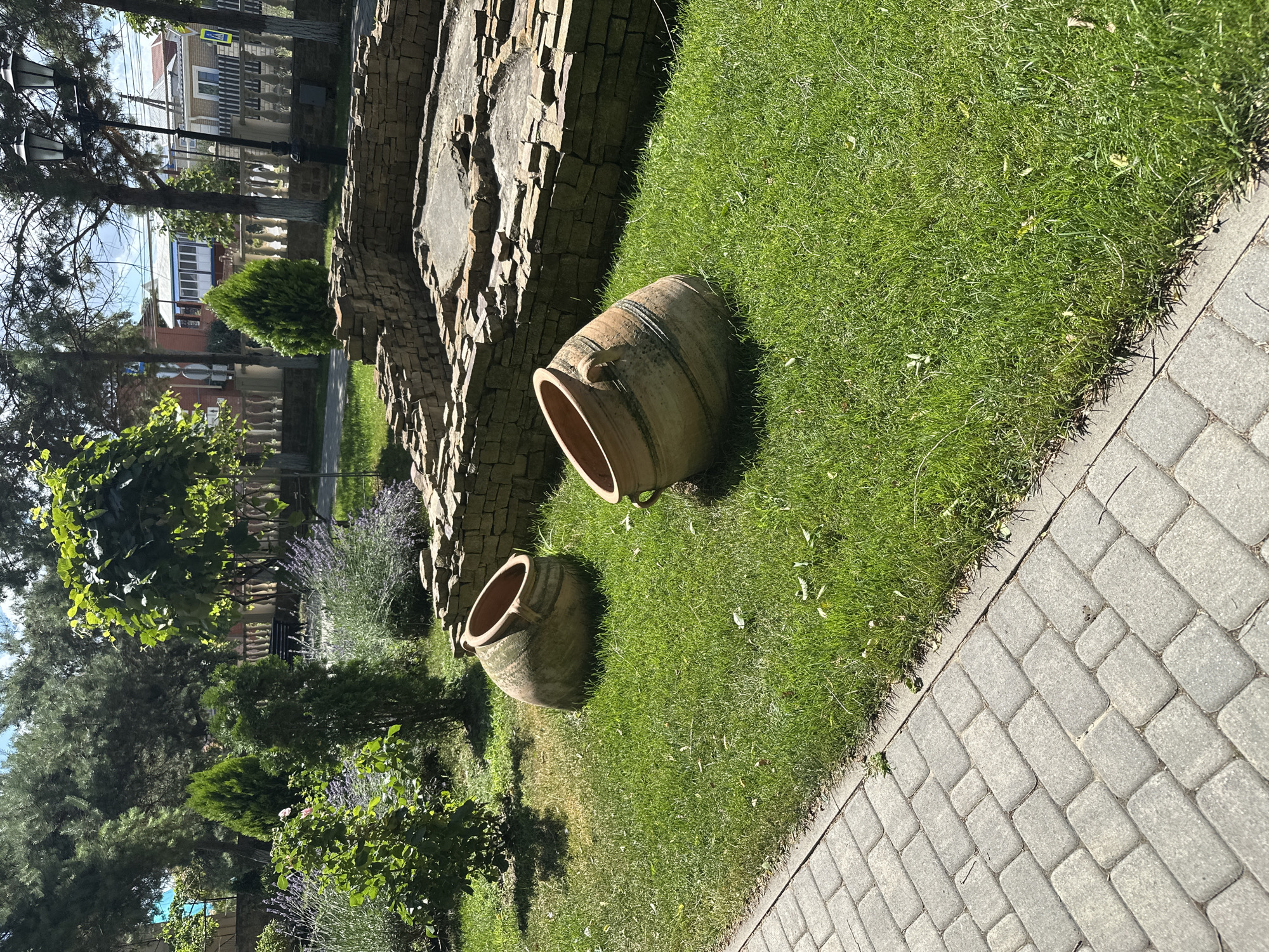 In Taman, Russia, an outdoor scene features a paved walkway bordering a lush green lawn. Two large, weathered ceramic amphorae, one positioned closer to the foreground and the other slightly further back, rest on the grass. Behind them, a low, rough stone wall encloses a sunlit, partially ruined structure with a concrete base and stacked stone foundations. The area is surrounded by various trees, including evergreens and leafy varieties, and in the background, residential buildings with tiled roofs are visible. The sky is bright and clear.