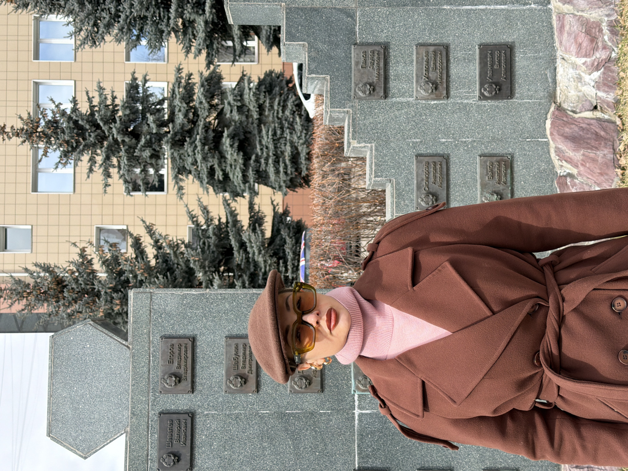 An outdoor, daytime portrait captures a stylish individual standing in front of a grey stone memorial monument in Krasnoyarsk, Russia. The person, positioned slightly to the right, is wearing a brown, belted trench coat with a prominent collar, lapels, and visible buttons. A light pink turtleneck sweater is worn underneath, complemented by a brown flat cap and distinctive olive-green framed sunglasses with orange-tinted lenses. Gold hoop earrings are also visible.
The large, tiered memorial monument is constructed from dark grey granite and features multiple bronze plaques, each engraved with a name and patronymic in Cyrillic script. Visible names on these plaques include "Шевелев Валерий Иванович," "Егоров Владимир Владимирович," "Шабунин Владимир Дмитриевич," "Холопьев Евгений Федорович," "Быков Анатолий Иванович," and "Иванкевич Виктор Алексеевич," among others, indicative of a commemorative purpose.
Behind the monument, several tall evergreen trees, likely firs or spruces, rise towards a light-colored, multi-story building with numerous windows. The ground around the monument includes a rough stone border and some sparse, dry vegetation, suggesting either late autumn or early spring. The bright lighting conditions indicate a clear, sunny day. The individual is posing for the camera, with a confident demeanor partially obscured by their sunglasses.