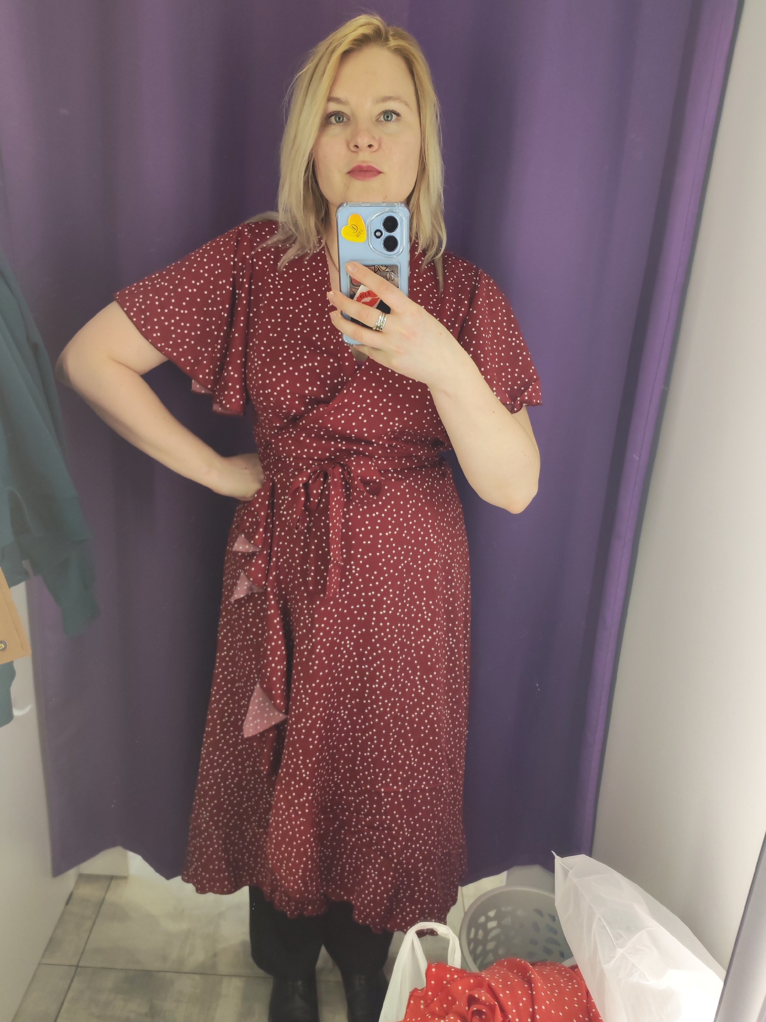 A blonde woman is taking a selfie in what appears to be a changing room, likely in Izhevsk, Russia. She is facing the camera, holding a light blue smartphone with yellow heart and other stickers in her right hand, positioned to capture her reflection in a mirror. She has light-colored eyes and red lipstick.
The woman is wearing a dark red midi-length dress with small white polka dots, featuring a V-neck, short flutter sleeves, and a tie-waist with some ruffles on the skirt. She has one hand on her hip. Black leggings or boots are visible beneath the dress. The background consists of a dark purple changing room curtain to her left, and the mirror reflects the white wall of the fitting room to her right. A clothes basket or bag with some red fabric (possibly part of the dress or another garment) is visible on the floor in the bottom right corner, alongside another white bag. The floor is tiled. No specific time of day or weather can be determined. There is no visible text.