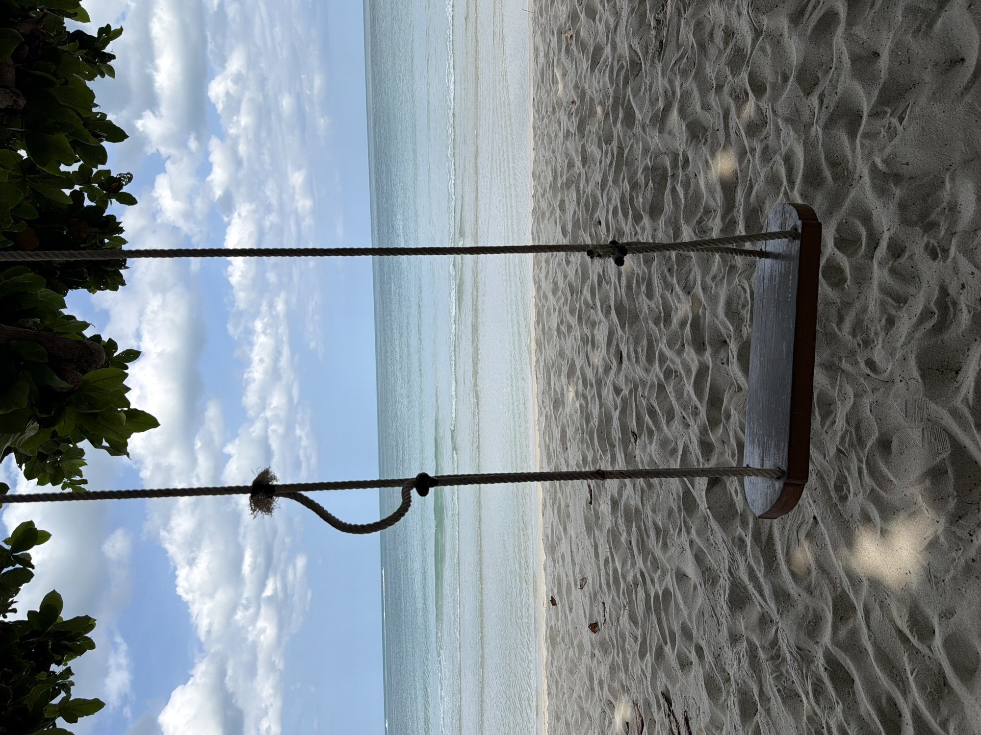 A wooden swing hangs from a tree branch over a sandy beach. The ocean is visible in the background, with gentle waves lapping at the shore. The sky is blue with scattered white clouds. The scene appears to be a tranquil and scenic spot, likely in a tropical location like Ko Samui District, Thailand, given the lush green foliage and the clear blue water. There are no people present in the image.