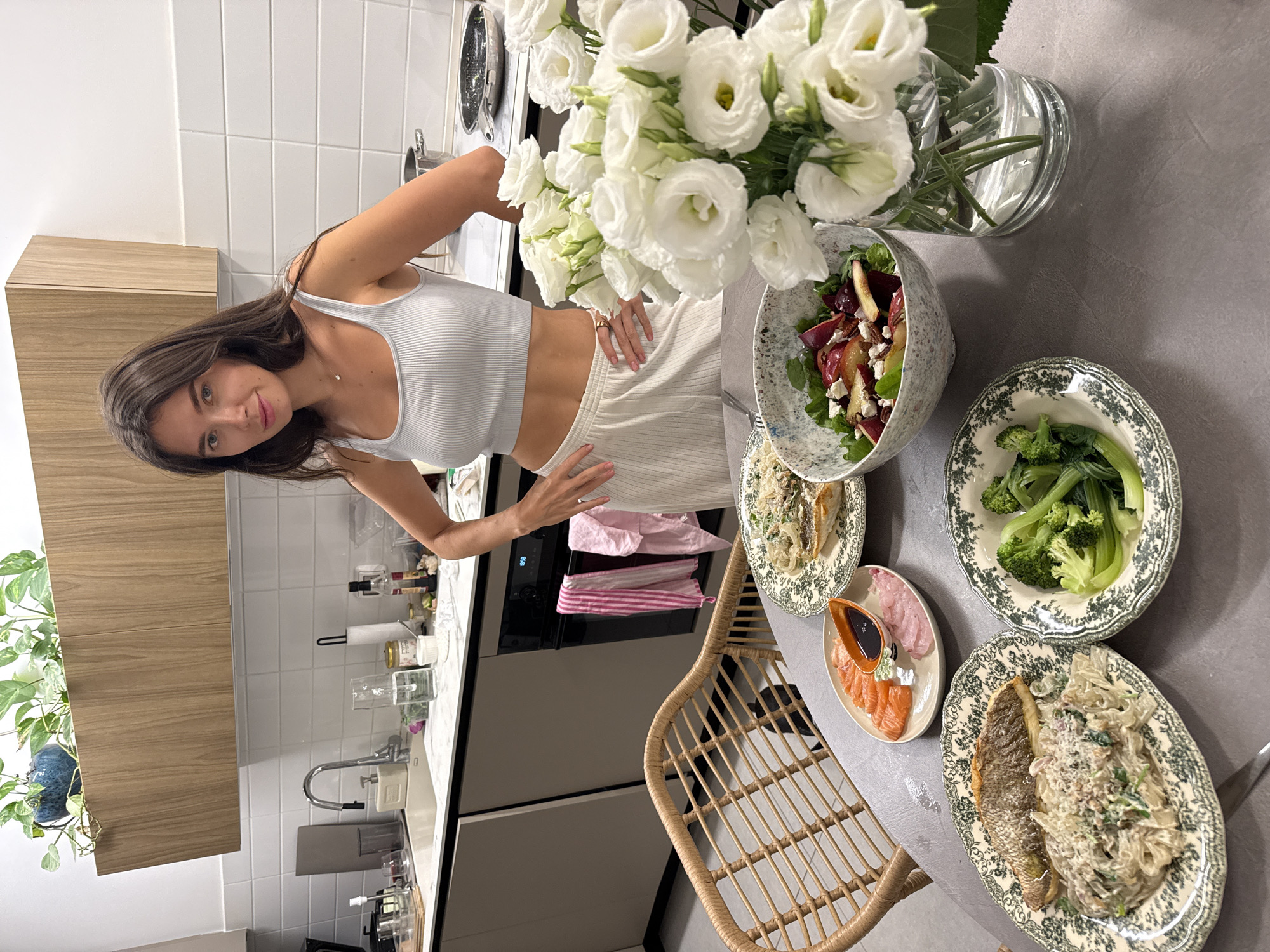 An indoor scene captures a young woman posing in a modern kitchen and dining area, rotated 90 degrees counter-clockwise from the original orientation. The woman, with long dark hair, stands with one hand on her hip, smiling gently towards the camera. She is wearing a white ribbed crop top and light-colored, possibly white, wide-leg pants, and a delicate necklace.
The foreground features a dining table laden with a variety of dishes. A large, clear glass vase holds an abundant bouquet of white Eustoma flowers, serving as a prominent centerpiece. The meal includes a generous bowl of salad containing mixed greens, red beets, and feta cheese. Also on the table are a plate of steamed broccoli, a decorative oval plate with what appears to be a whole baked fish (possibly sea bream) accompanied by creamy pasta, and a smaller oval plate with slices of raw salmon (sashimi) and another type of fish, alongside a dish of soy sauce. The food is presented on ornate green and white patterned plates. A wicker chair is partially visible beside the table, suggesting a meal setting.
In the background, a contemporary kitchen is visible with white subway tile backsplash, dark grey lower cabinets, and light wooden upper cabinets. A stainless steel sink with a faucet, various kitchen utensils, jars, and a soap dispenser are on the counter. An oven with pink and white striped towels hanging from its handle is also seen. A potted plant with lush green leaves sits on a shelf, adding a touch of greenery to the space. The lighting suggests an indoor setting, likely evening or late afternoon. No visible text is present. The overall ambiance is clean, modern, and inviting, centered around a prepared meal.