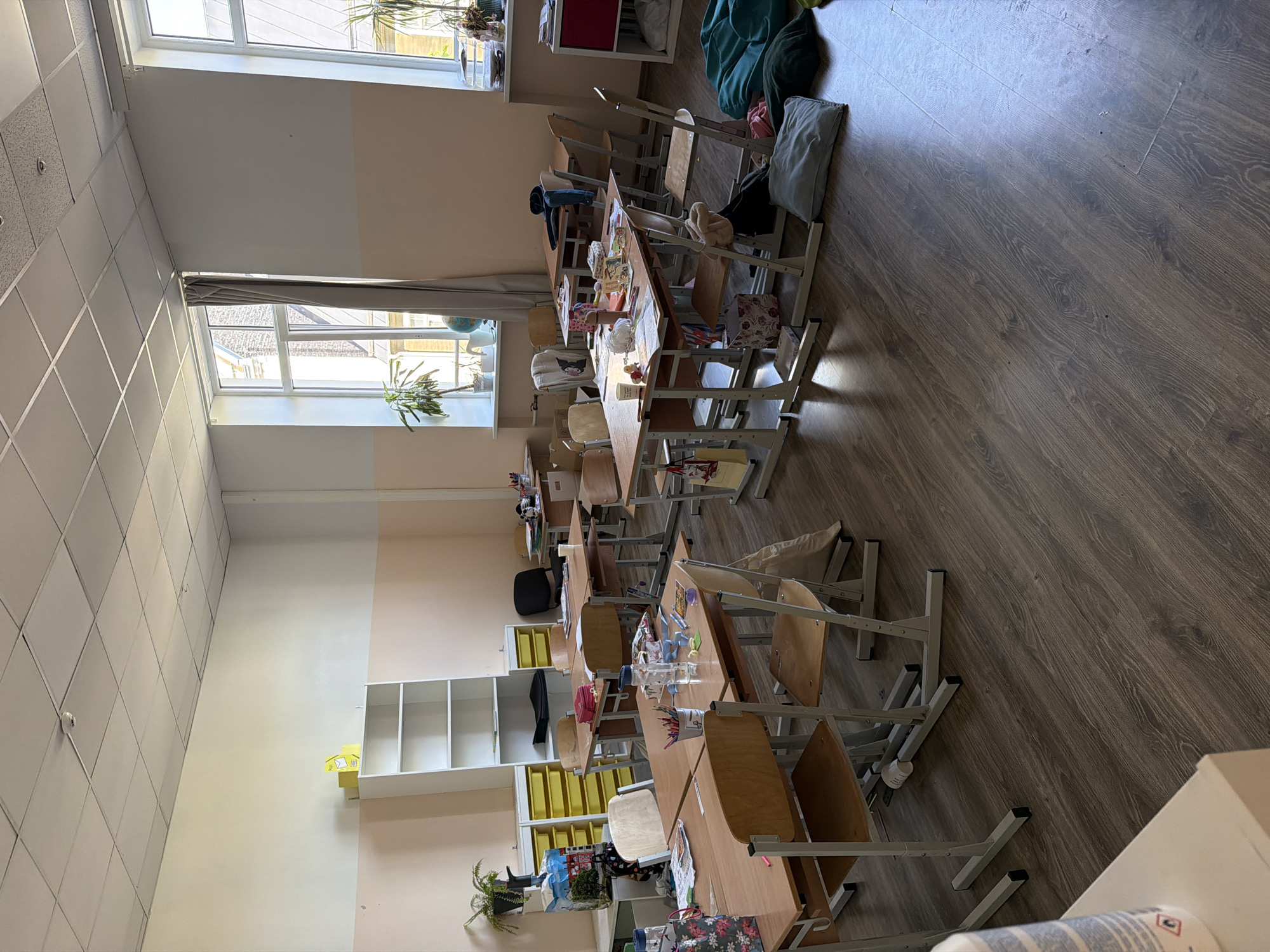 The image depicts a classroom with rows of desks and chairs. The setting appears to be a school, likely in Riga, Latvia, given the provided context. Sunlight streams in from large windows, suggesting it is daytime. The room has a warm, neutral color palette with light-colored walls and a wooden floor.
The desks are arranged in pairs, with some chairs positioned at them and others pushed back or pulled out. Many desks are cluttered with various objects, including books, papers, colorful containers, and decorative items like stuffed animals and small boxes. This suggests either a recent activity or that the room is in a state of everyday use, perhaps between classes or at the end of the school day.
There are no people visible in the image, but the presence of student belongings implies their recent or anticipated presence. The overall atmosphere is one of a functional learning environment, but with a touch of personal items and a general sense of tidiness, though not perfectly ordered.
Notable details include the ceiling tiles, which are a mix of square and diamond patterns, and the window frames, which are white. A tall shelving unit with cubbies is visible against one wall, and a yellow organizer filled with trays is placed on the floor next to it. In the background, several large beanbag chairs are present, adding a casual element to the classroom. A plant is visible on a windowsill. There is no visible text in the image.