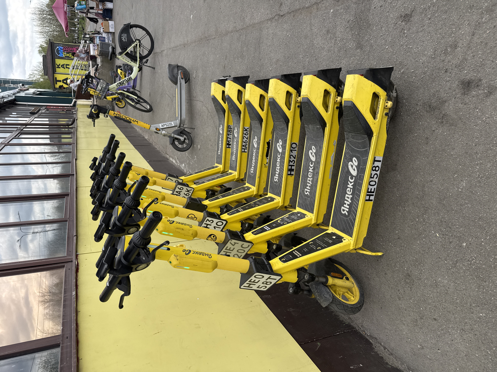 A row of yellow electric scooters with "Yandex Go" branding are lined up on a paved surface in Saint Petersburg, Russia. Adjacent to the scooters is a yellow building with window panes. Further in the background, there is an assemblage of bicycles, a canopy, and an outdoor vendor stall. The scooters have license plates on the rear. The image is taken in daylight.