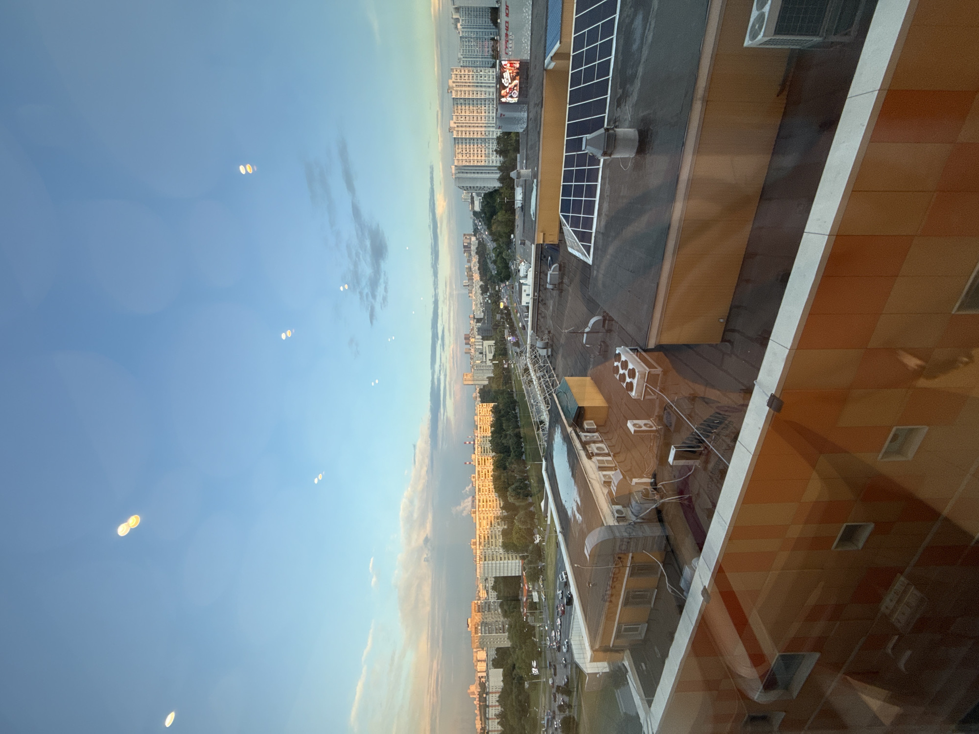 This is a high-angle, wide shot of a city skyline at dusk, taken from a window. In the foreground, the top of a modern building with orange and yellow tiled walls is visible, reflecting the sky. Beyond this building, the roofs of other structures are visible, including one with solar panels and several air conditioning units.
In the mid-ground, a street with cars is visible, lined with trees. Further in the distance, a dense urban landscape unfolds, featuring a variety of apartment buildings of different heights and architectural styles. Some of these buildings are bathed in the warm, golden light of the setting sun, creating a contrast with the cooler blue and pink hues in the sky. Smoke or steam is rising from several industrial chimneys in the distance.
The sky above is a gradient of blue, with scattered clouds tinged with pink and orange. There are several circular light refractions, or bokeh, visible in the sky, indicating they are likely from lights inside the building where the photo was taken.
While the image depicts an urban environment in Moscow, Russia, specific landmarks are not clearly identifiable without further information. The scene suggests a residential and commercial district, characterized by its multi-story buildings and infrastructure. There are no discernible people or animals in the image. The overall impression is one of a bustling city settling into the evening.