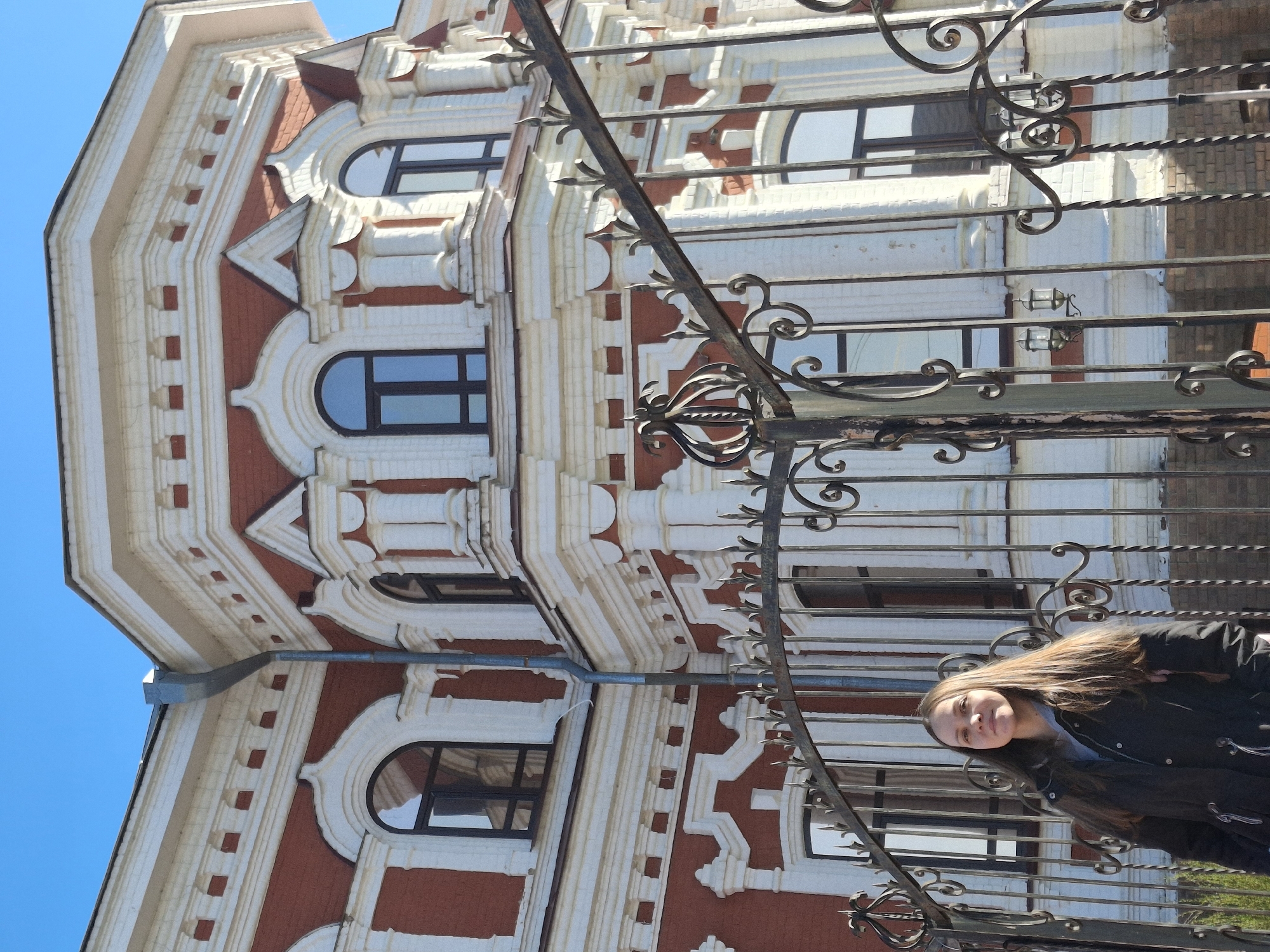 A young woman with long brown hair stands behind a wrought iron fence in Sarapul, Russia. She wears a black jacket and smiles slightly at the camera. Behind her is an ornate three-story building with red brick and white decorative trim, arched windows, and a blue sky in the background. The image is rotated counter-clockwise.