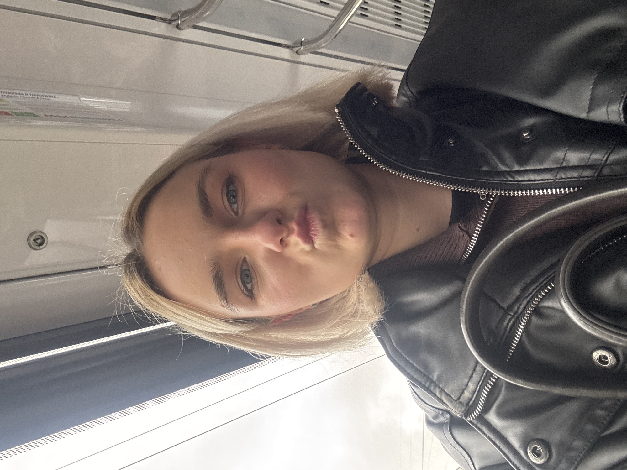 A selfie taken in what appears to be a train car of the Moscow Metro in Russia. A young, fair-skinned woman with blonde, shoulder-length hair is the primary subject. Her lips are pursed, indicating a possible attempt at a kiss. She is wearing a black leather jacket over a brown sweater. The background shows the typical interior of a Moscow Metro train car, including the window, the wall, and the handrails. The lighting is slightly dim, likely due to being indoors.