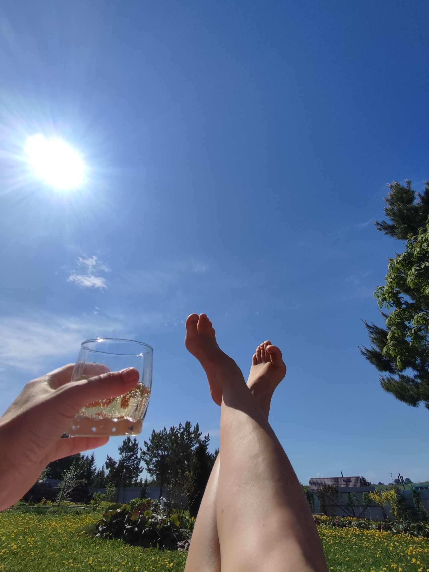 A first-person, eye-level shot of a sunny outdoor scene. The sky is bright blue with a few scattered clouds and the sun shining brightly in the upper left corner. In the foreground, a person is holding a glass filled with a light-colored liquid, possibly a sparkling beverage. The person's crossed legs extend upwards towards the center of the frame, ending with bare feet. The background features a green lawn dotted with yellow wildflowers, various trees, and glimpses of houses, indicating a residential area. The location appears to be Фоки, Russia.