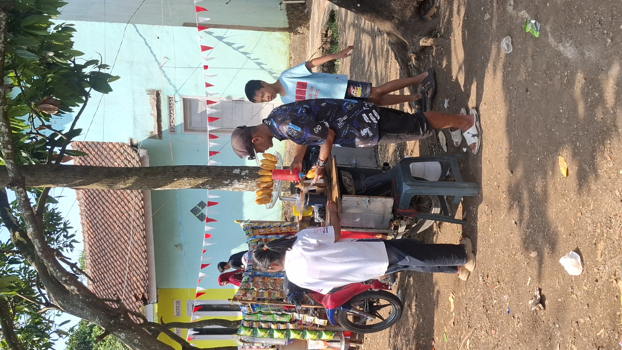 In Cilaku, Indonesia, a street vendor prepares corn dogs at a cart set up outdoors. The cart, attached to a red and blue motorcycle, is laden with various bagged snacks. A man wearing a dark baseball cap and a patterned shirt is seen dispensing sauce onto a corn dog. Next to him, a young boy in a light blue t-shirt stands observing. A young woman in a white shirt and dark pants is also present, seemingly assisting or waiting. In the background, two houses with red tiled roofs are visible, decorated with red and white triangular flags. The ground is a mixture of dirt and scattered leaves, with some litter present. The scene is illuminated by daylight, suggesting an outdoor market or roadside stall.