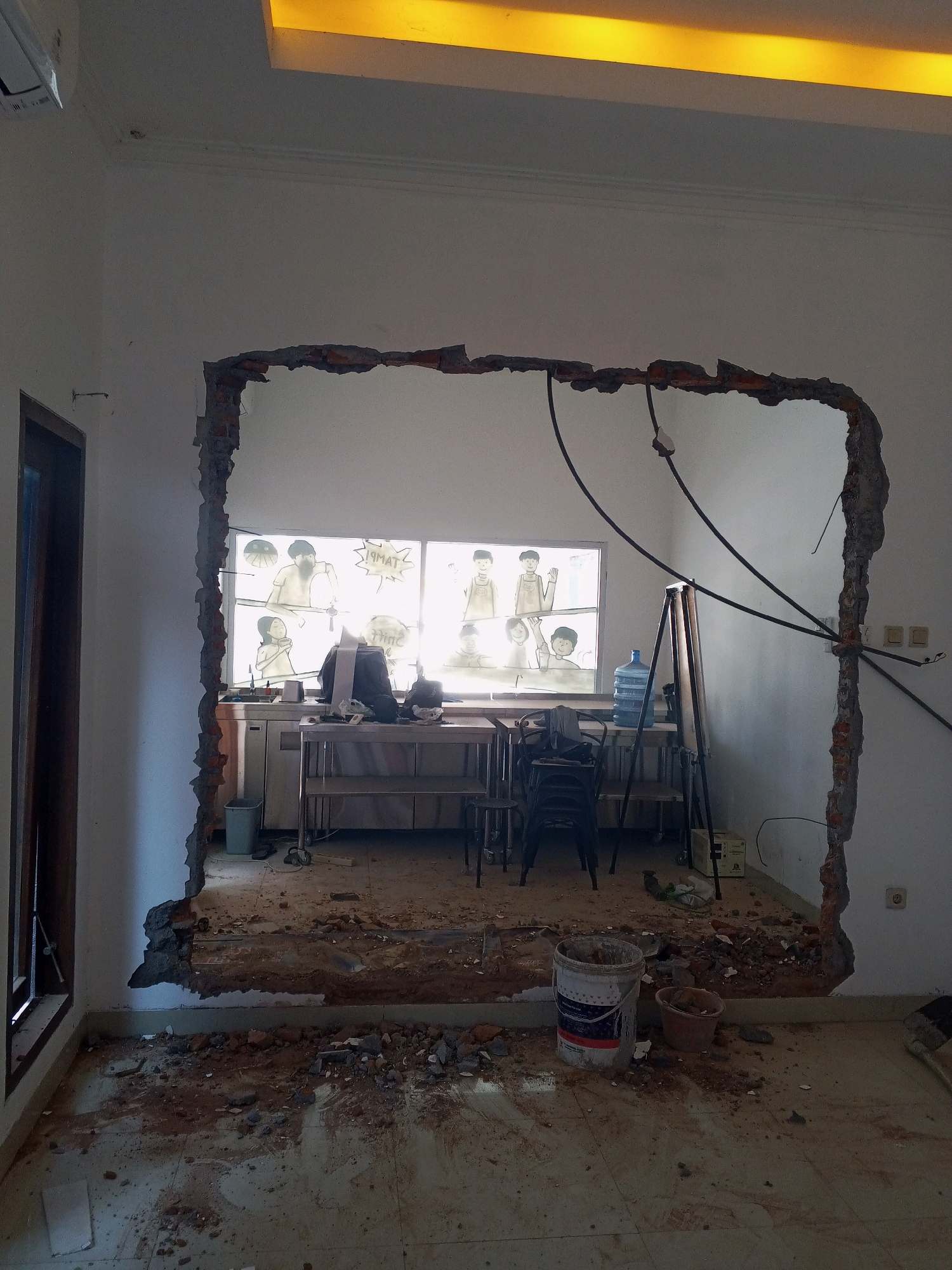 The image shows a room under renovation in Condongcatur, Indonesia. A large section of a wall has been demolished, creating an opening. The floor is covered in debris from the demolition, including bricks and dirt. In the background, through the opening, a window is visible with cartoon-like drawings on it, possibly from a previous decoration. There is furniture visible behind the opening, including a counter, chairs, and a water cooler. A bucket containing some material, possibly for construction or cleaning, is in the foreground. A light fixture with a yellow glow is visible at the top of the image. Electrical wires are also exposed from the demolished wall. The overall scene suggests an ongoing construction or renovation project.