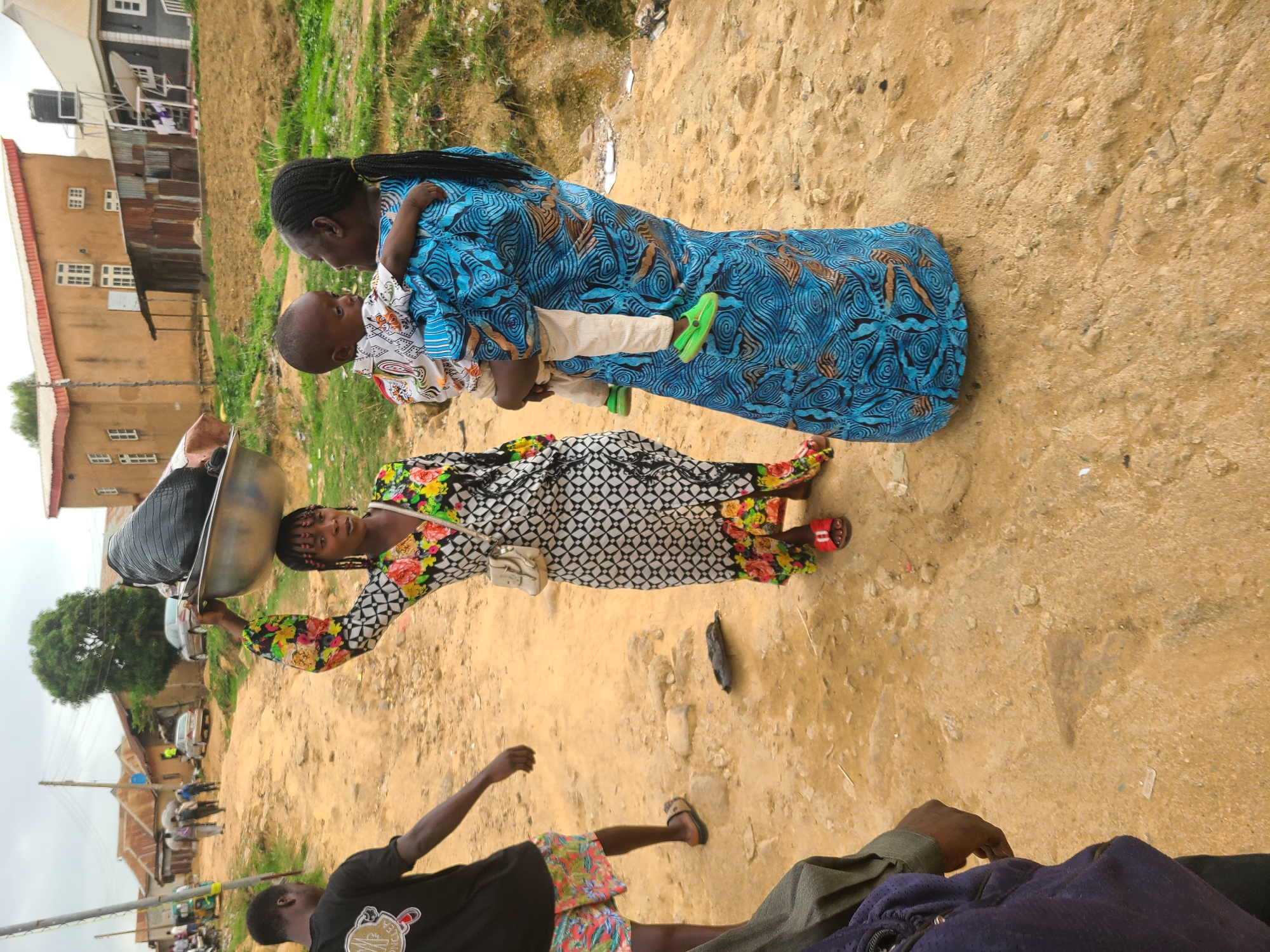A street scene in Unknown, Unknown features two women. One woman is carrying a large metal bowl of items on her head. She is wearing a long-sleeved floral top, a black and white geometric print dress, and red sandals. The other woman is carrying a baby. She is wearing a long blue dress with a brown and blue pattern. The baby is wearing a patterned shirt and light-colored pants and green sandals. In the background, there are buildings and people visible. The ground is dirt. In the foreground, there are two men in casual clothing.