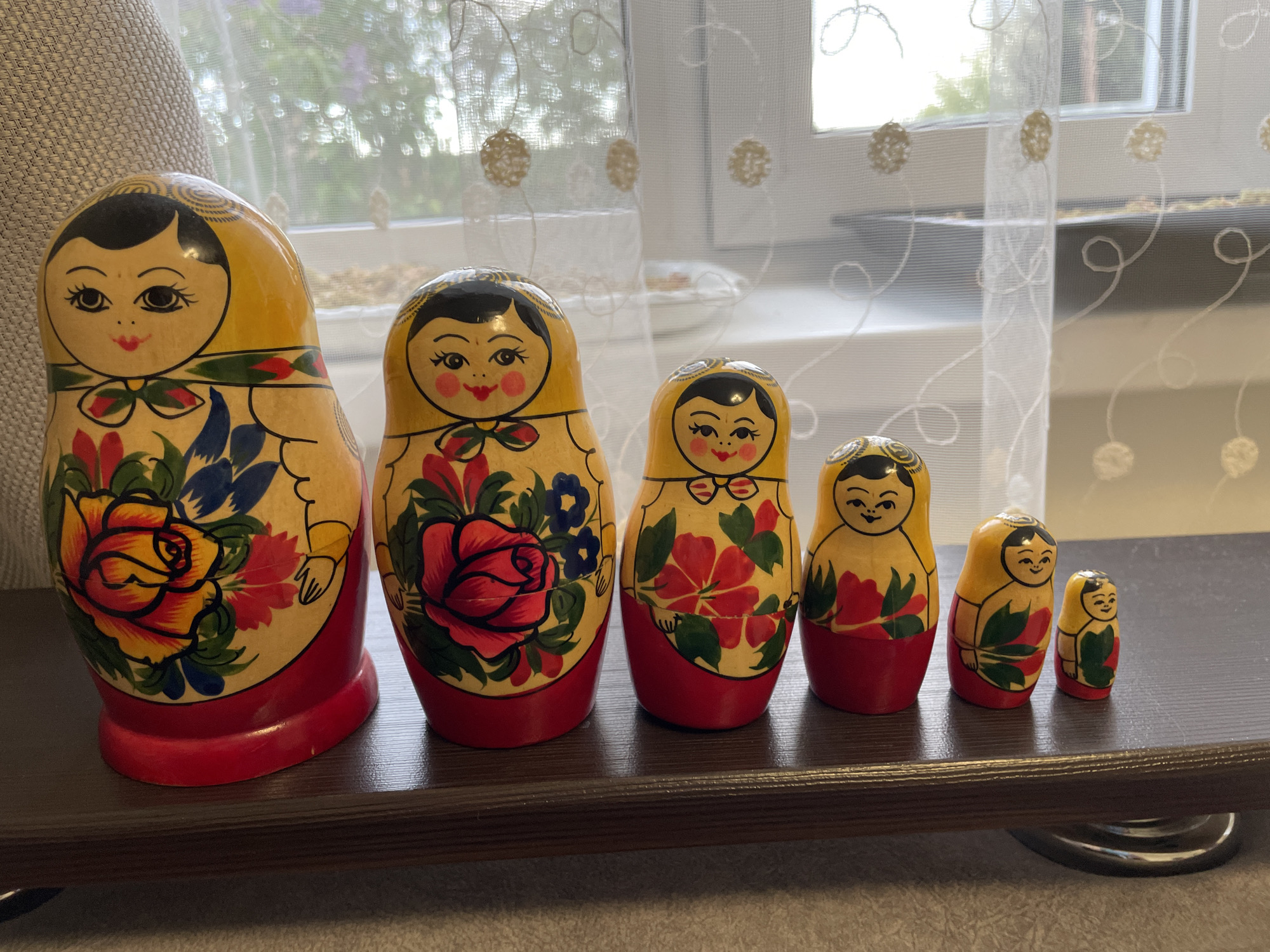 A photograph shows a collection of six Matryoshka dolls, also known as Russian nesting dolls, arranged in a line from largest to smallest. The dolls are painted with floral designs, predominantly featuring roses, and have red bases. The dolls are set on a dark brown surface, which appears to be a shelf. In the background, a window is covered with a sheer, patterned curtain. The location is not evident from the image.