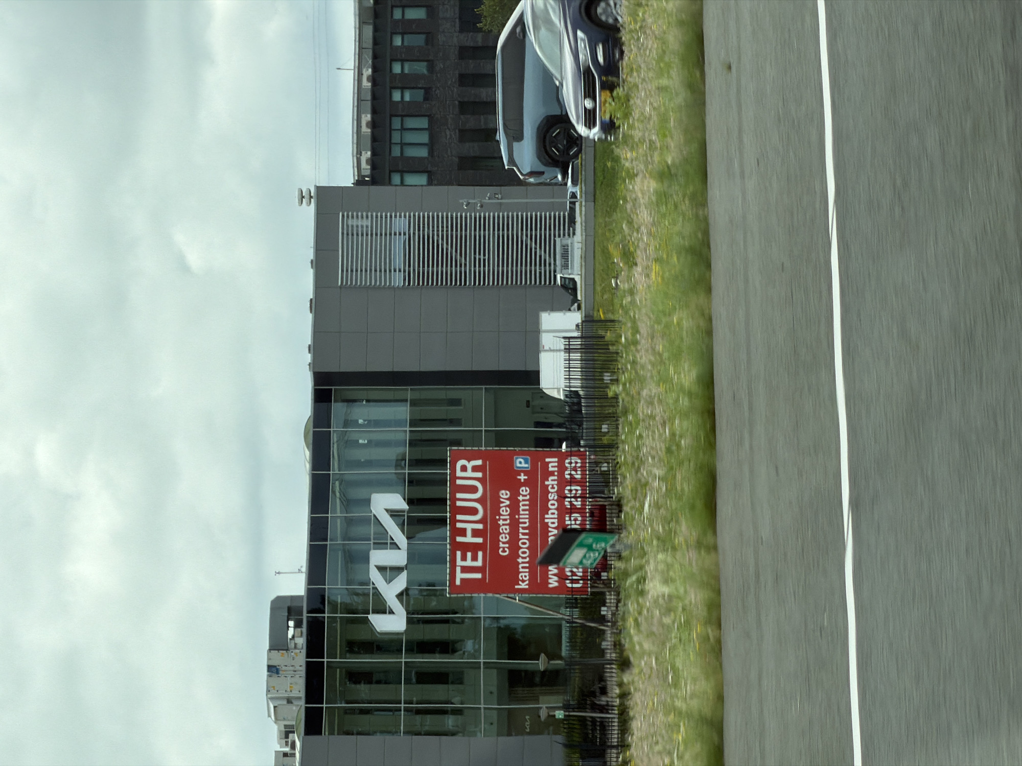 The image shows a modern building with a large "KIA" logo visible on its glass facade. In front of the building, a red sign with white text reads "TE HUUR" (For Rent) and advertises "creatieve kantoorruimte + P" (creative office space + parking). Below this, a website "vdbosch.nl" and a phone number "0275 29 29" are displayed. A dark blue SUV is parked in front of the building to the right of the sign. The scene appears to be a commercial area with a road in the foreground, suggesting it was taken from a vehicle. The sky is overcast. The location is Nieuwer Ter Aa, Netherlands, as indicated by the specific details on the sign and the overall architectural style.