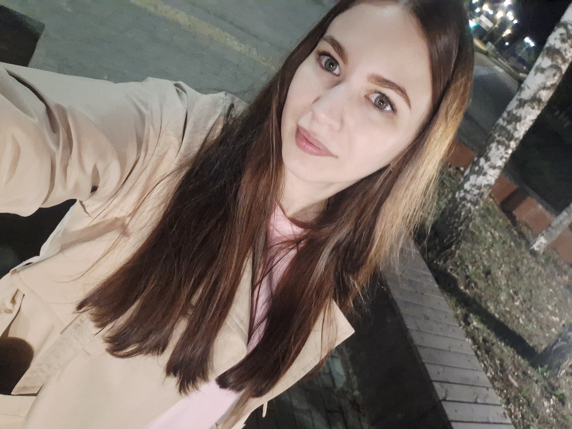A woman with long brown hair, fair skin, and green eyes is taking a selfie outdoors at night in what appears to be a park or public space in Chaikovsky, Russia. She is wearing a light pink shirt and a beige trench coat. The background features a birch tree with white bark, wooden boardwalk, dimly lit street lights, and foliage. The image has a slightly grainy quality, suggesting it was taken in low light conditions.
