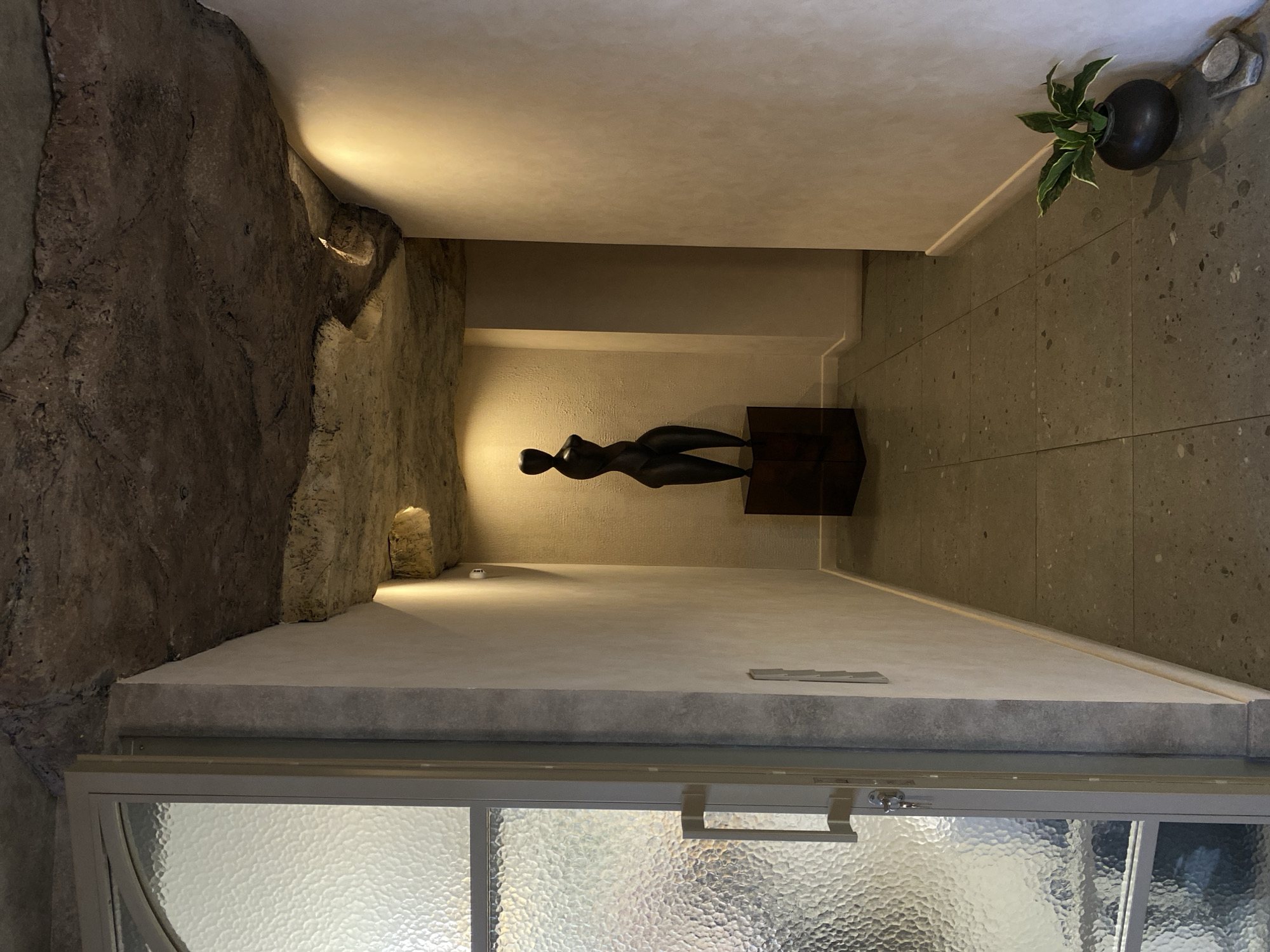 The image shows an interior space with a modernist aesthetic. A human figure sculpture sits on a dark wooden plinth. The sculpture is placed in a recessed niche. The walls are a blend of natural rock and smooth, light-colored plaster or concrete. The flooring is tiled. A plant is present in a dark, round pot. A window with textured glass is visible in the foreground. The location in Izhevsk, Russia is not verifiable from the visual information.