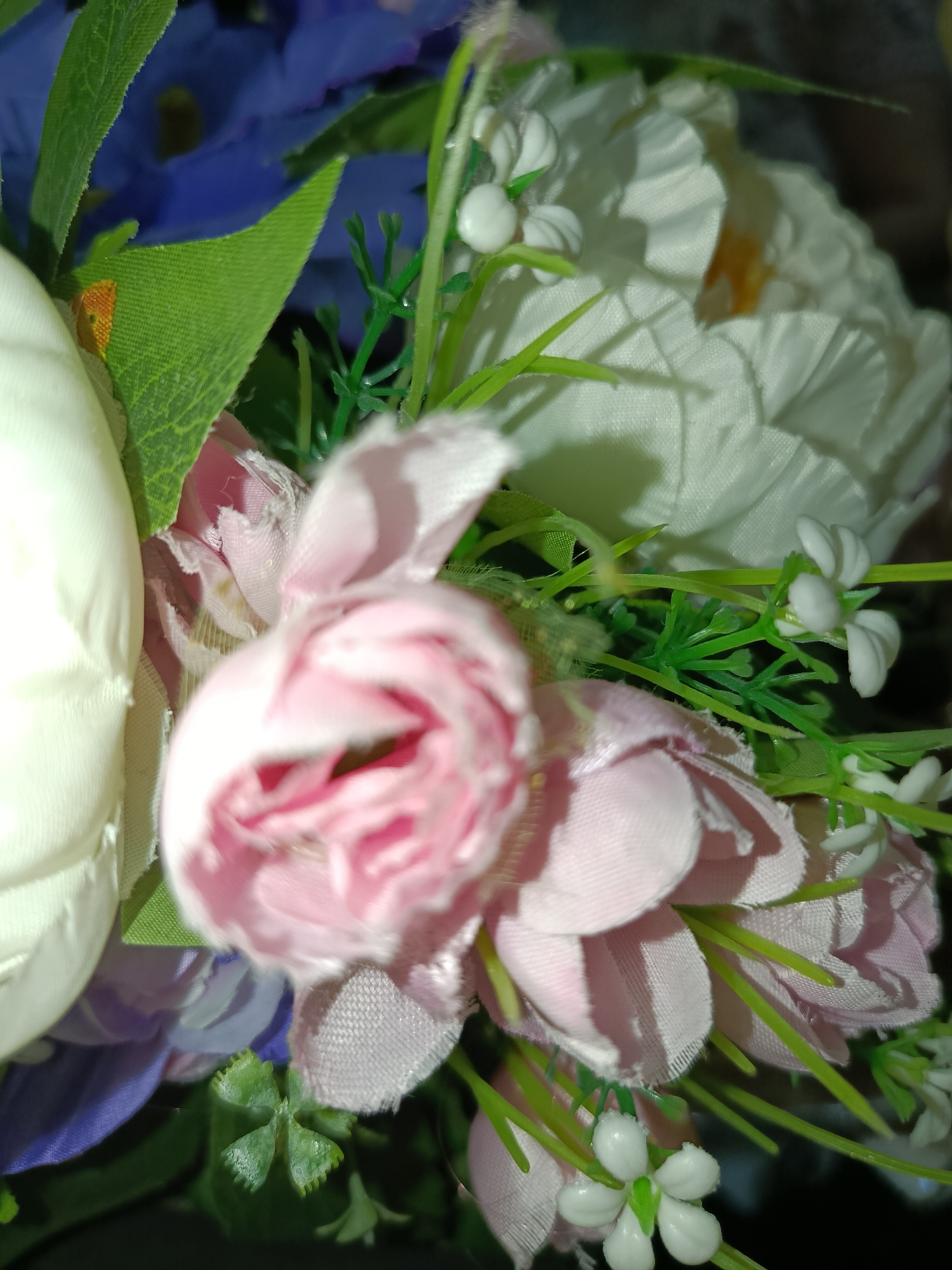 The image is a close-up, blurred shot of artificial flowers. The flowers are a mix of pink roses, white flowers, and some blue and purple flowers in the background. There are also green leaves and stems visible. The lighting is dim, with some harsh shadows cast on the petals. The focus is on the pink rose in the foreground, which is soft and out of focus. The background is even more blurred, making it difficult to discern any specific details. It is not possible to determine the location in Tashkent, Uzbekistan, or any actions or interactions, as the image only contains inanimate objects.