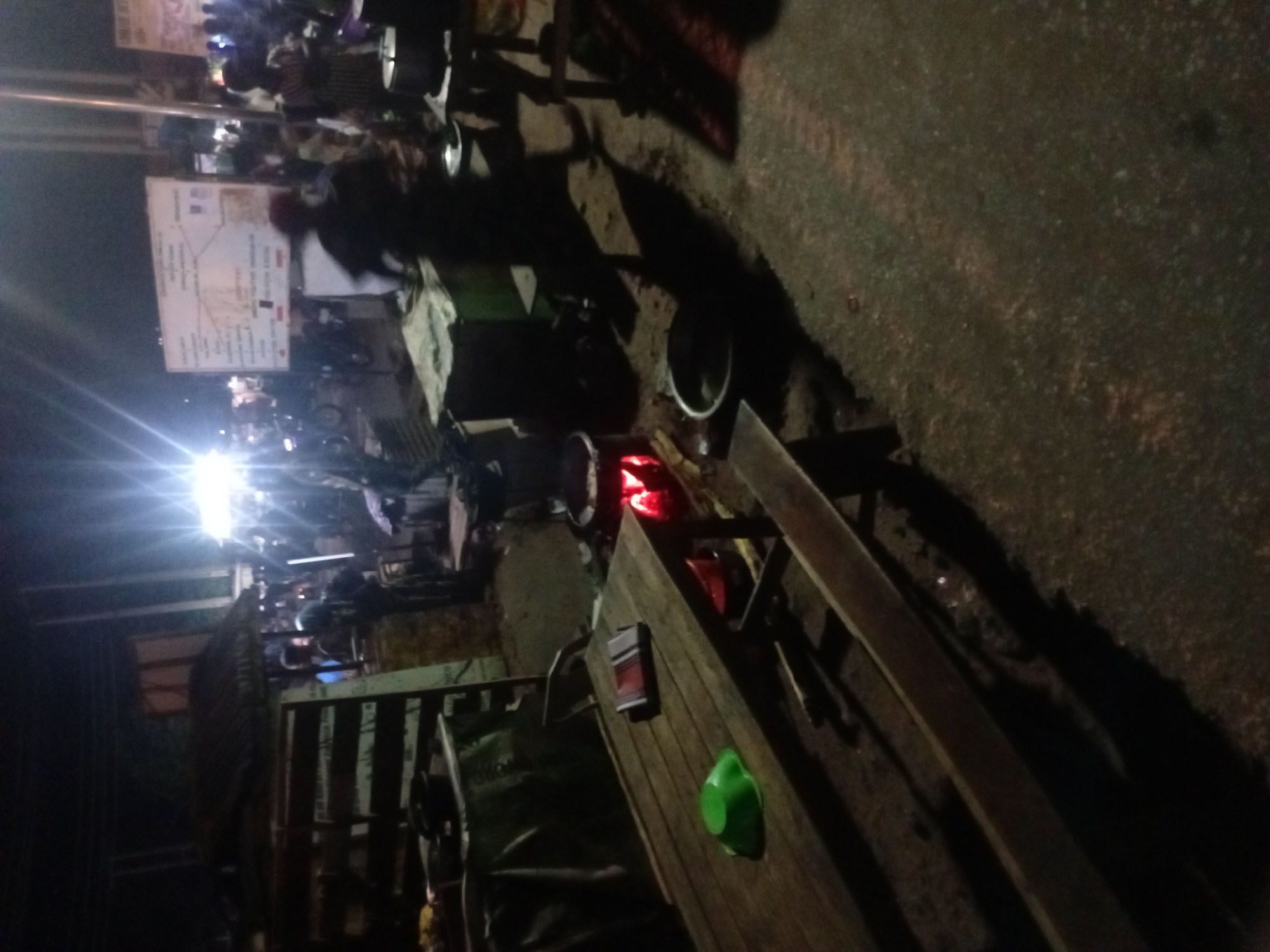 The image depicts an outdoor scene in Lokoja, Nigeria, at night. A brightly lit area is visible in the background where a large crowd is gathered around what appears to be a map or informational sign. In the foreground, a wooden table and bench are present, with a green object resting on the table. A cooking setup with a pot and visible flames suggests food preparation or street vending. The overall environment seems to be a roadside area with limited lighting. The image is taken at an angle, distorting the perspective.