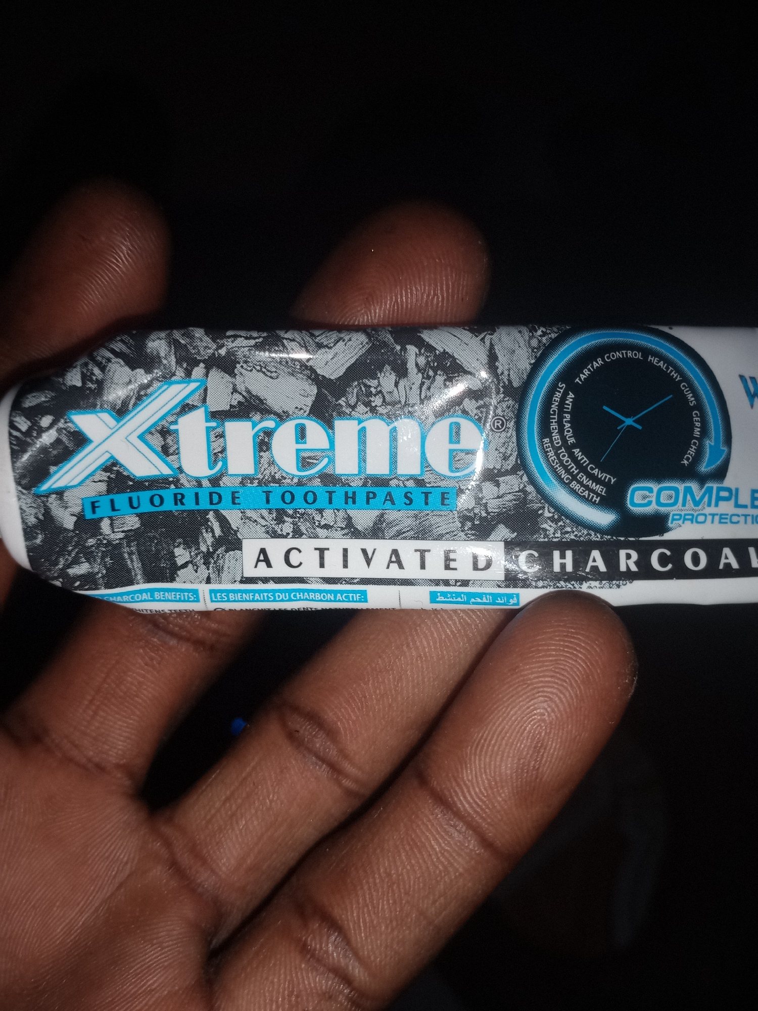 The image shows a close-up view of a tube of "Xtreme" brand fluoride toothpaste with activated charcoal. The tube has a black and gray patterned design with white and blue text. The words "Xtreme" and "FLUORIDE TOOTHPASTE" are prominently displayed. Below this, "ACTIVATED CHARCOAL" is written. There is also a circular graphic with text in English and potentially Arabic, indicating benefits like "TARTAR CONTROL," "HEALTHY GUMS," and "REFRESHING BREATH." The tube is held by a hand with dark skin, the fingers are visible in the foreground, obscuring parts of the product. The background is dark, suggesting an indoor or nighttime setting. There is no information in the image that indicates a specific location in the city of Jalingo, Nigeria, or any other geographical context.
