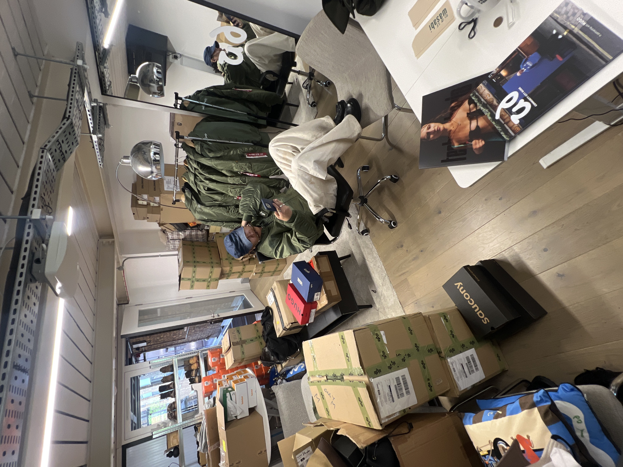 The image presents an indoor scene, likely a busy office or storage space in London, United Kingdom. A person is seated on an office chair, leaning back in a relaxed posture while looking at their smartphone. They are wearing an olive green jacket, light-colored trousers, and a blue cap, with their reflection visible in a large mirror on the left wall.
The room is characterized by a significant amount of merchandise and packaging. Numerous cardboard boxes are stacked on the light wooden floor and on metal shelves. Visible brand names on these boxes include "Saucony," "HUGO," and "VANS," along with the "Alpha Industries" logo on several packages. A clothing rack in the center holds multiple olive green bomber jackets.
On a white table to the right, an open magazine or book displays the text "COMMUNITY CITY GUIDE," "p2," and "HUGO," alongside a photograph of a woman. Other items on the table include scissors and a white mug with "wasabi" written on it. The space is well-lit by bright overhead fluorescent lights and natural light filtering in from a window or glass door in the background. Exposed metal shelving and electrical conduits contribute to an industrial aesthetic. The scene depicts a moment of relaxation or a break within a cluttered work environment, possibly related to inventory management or retail preparation. Additional visible text includes "24."