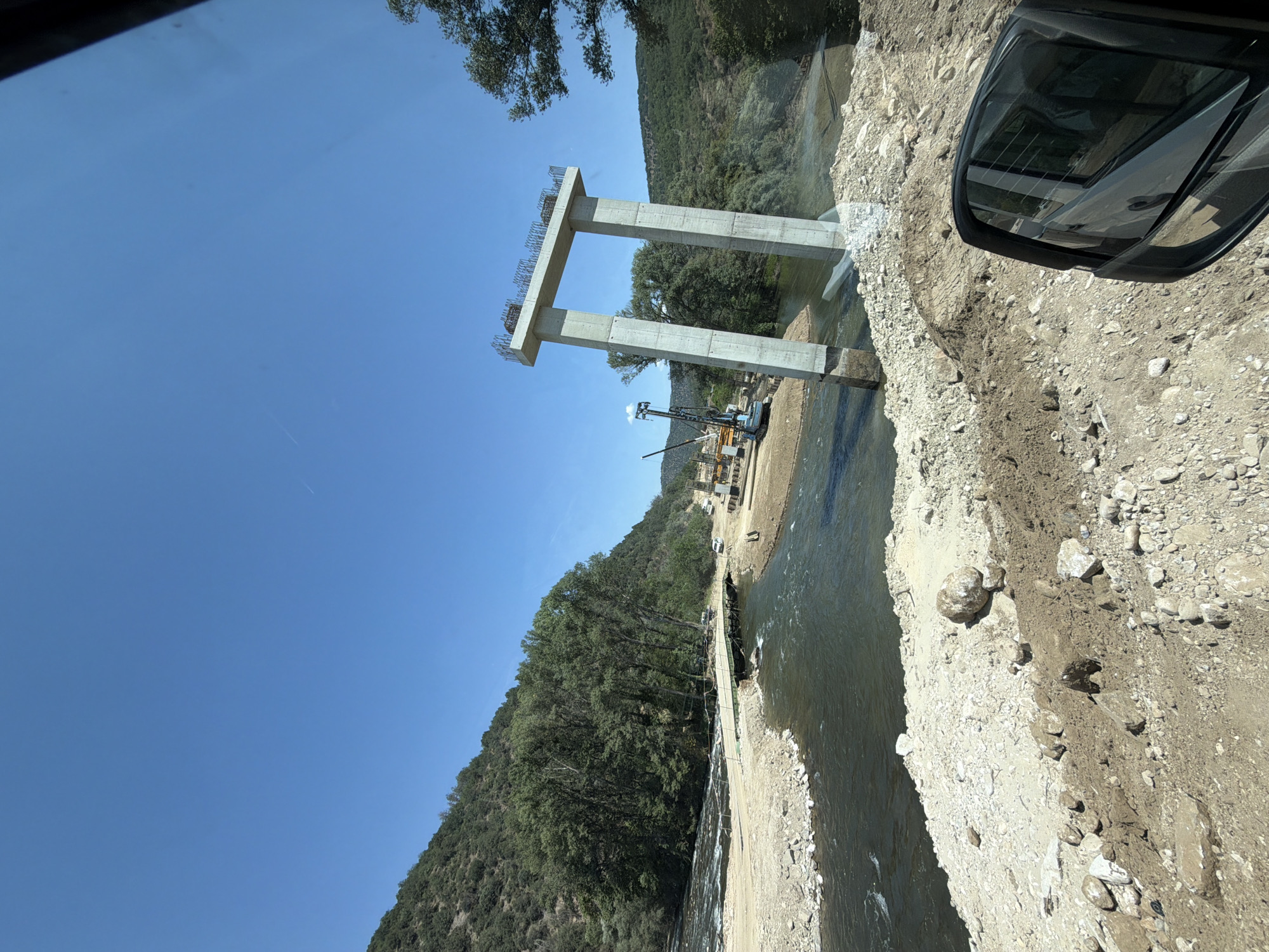 In the city of Kresna, Bulgaria, a bridge is under construction over a river. The scene is characterized by a bright, clear blue sky and lush green trees on the surrounding hillsides. In the foreground, a partially constructed concrete bridge support stands tall, resembling an archway with two pillars. On the left side of the river, a temporary bridge is in place, with several cars visible. A construction crane and other heavy machinery are positioned on the riverbank, indicating ongoing work. The river itself appears to be flowing moderately. The surrounding terrain is rocky and somewhat arid, with visible construction materials and earthwork. The image captures a moment of progress in infrastructure development amidst a natural landscape.
