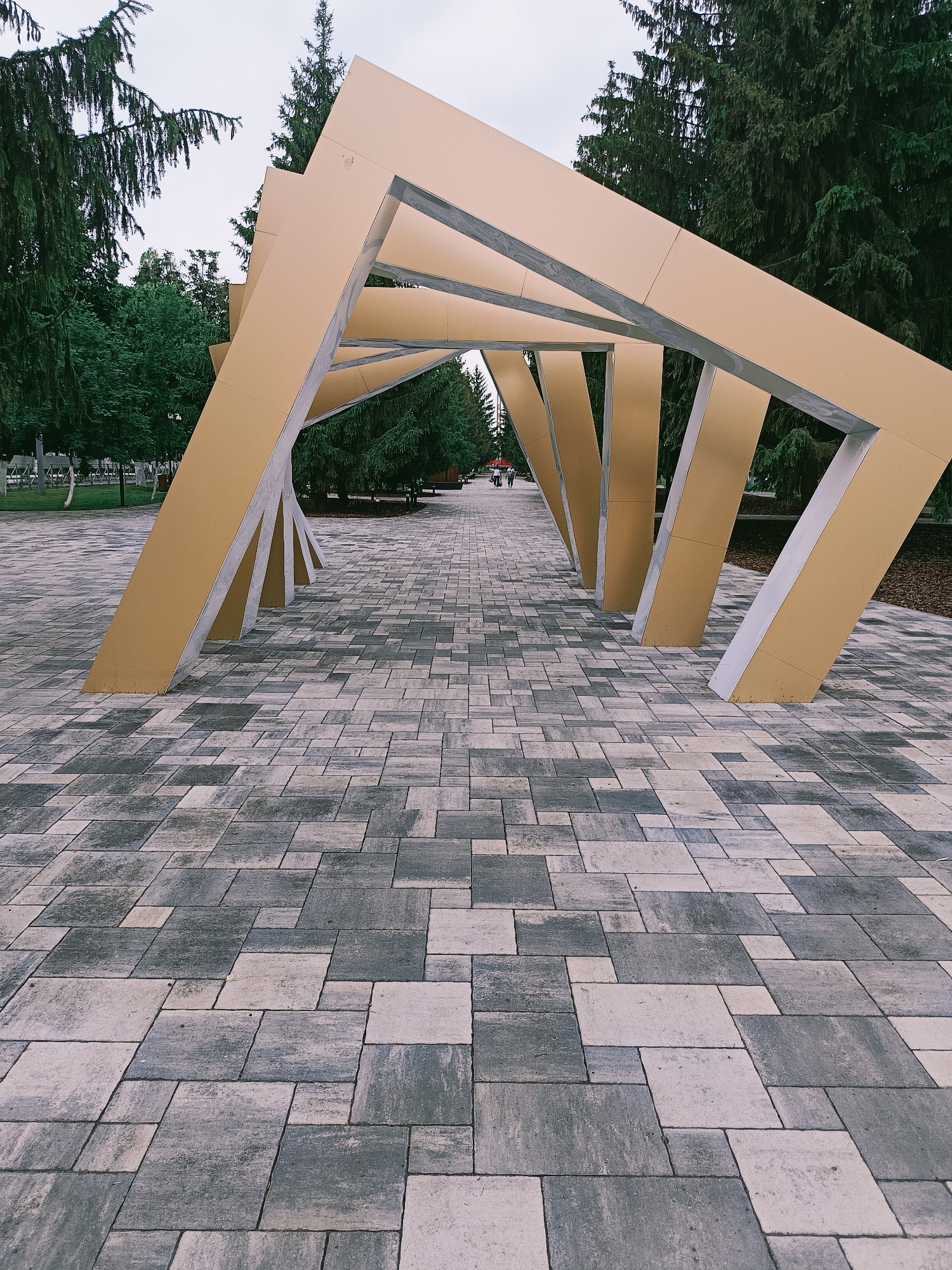 An outdoor, eye-level, full shot captures a decorative modern archway in a public park in Gubkin, Russia. The archway is made of golden-colored geometric panels supported by grey concrete pillars. The ground in front of the archway is paved with rectangular stones in varying shades of grey and beige, arranged in a staggered pattern. Tall, dark green coniferous trees line the sides of the pathway extending from the archway into the distance. The sky is overcast and light grey. Two figures, a man and a woman, are walking away from the camera on the pathway in the mid-ground, and there are a few more figures further in the distance.