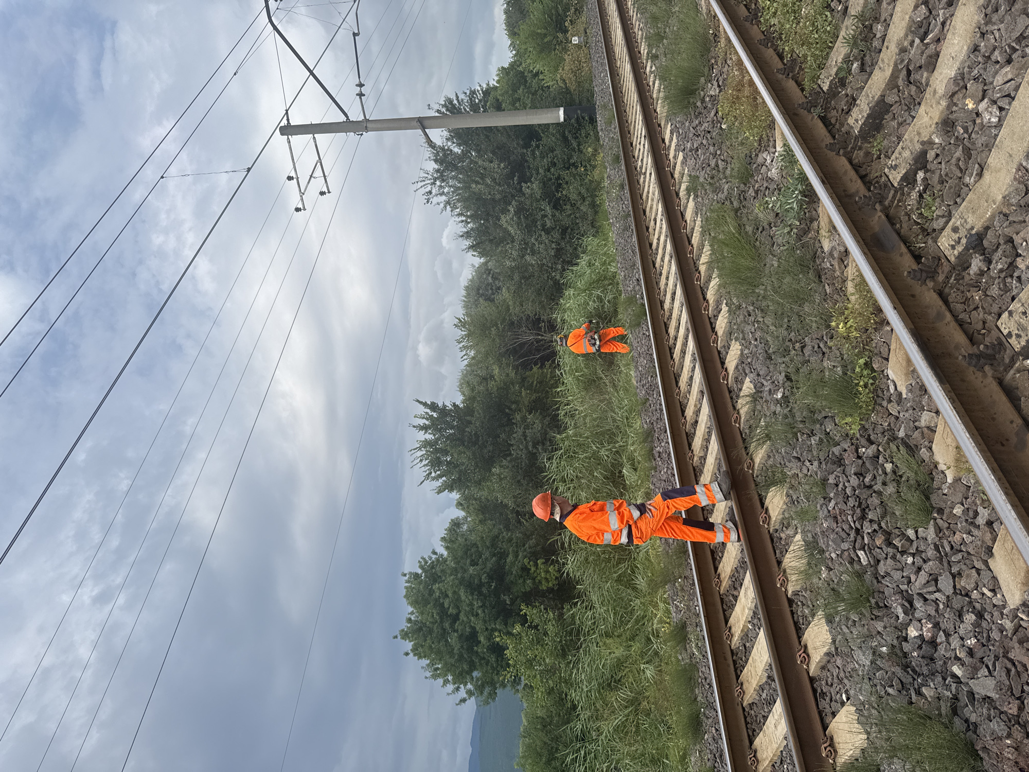 The image shows two individuals in high-visibility orange work attire on a railroad track. The individual in the foreground is walking on the tracks, carrying a tool. The individual in the background is using a string trimmer to clear vegetation. The scene is set in a rural area, with lush green trees and vegetation lining the tracks. Overhead, power lines for the electric railway are visible against a cloudy sky. The location is Kvemo Khvedureti, Georgia.