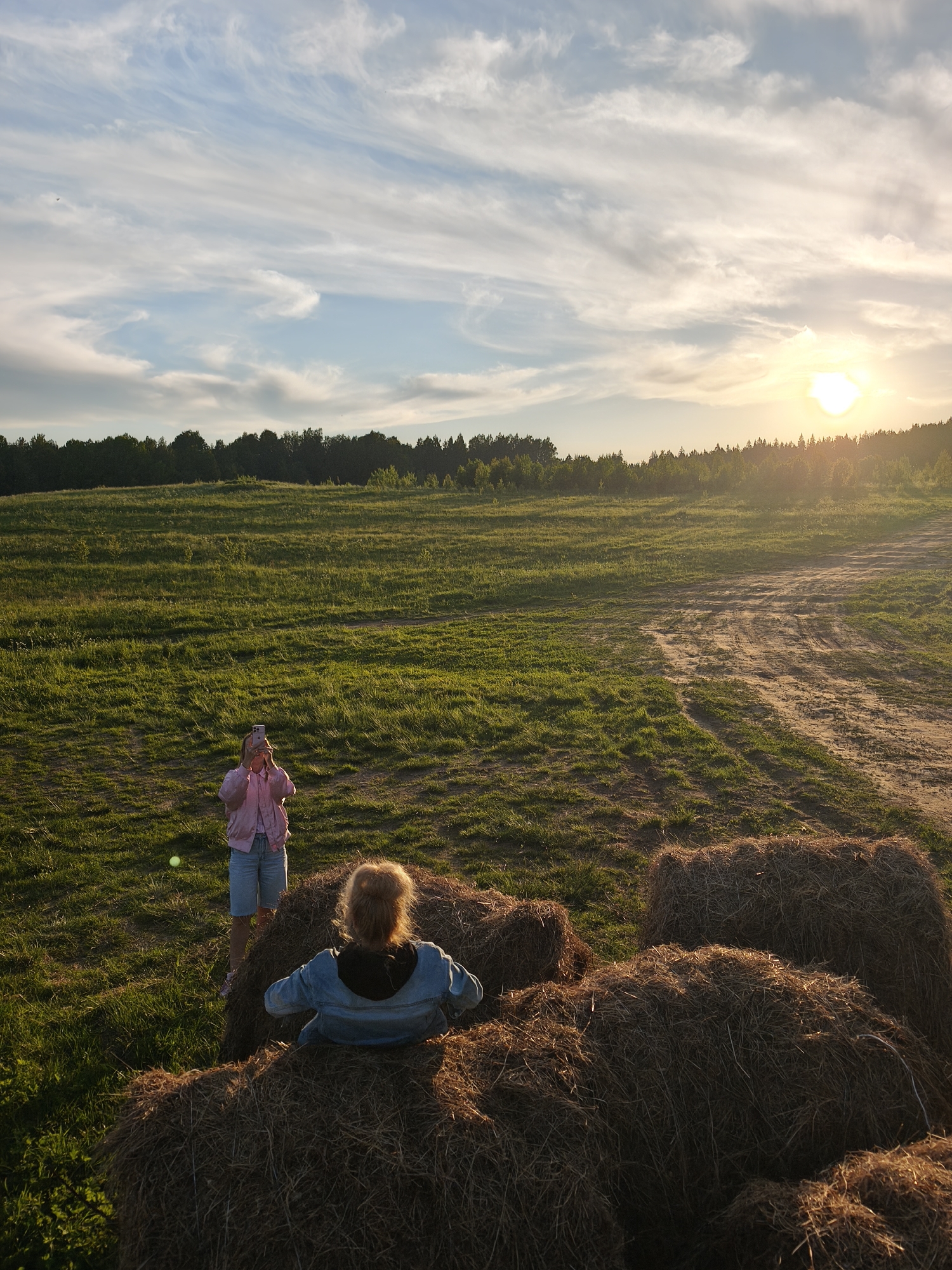 This image depicts a serene, sunlit rural landscape, likely in Стеклянный, Russia, during the golden hour of late afternoon or early evening. The scene is dominated by a vast, green, undulating field, bordered by a dense line of dark forest trees in the background. A dirt path or tracks are visible winding through the grass on the right side of the field.
In the foreground, several large hay bales are present. One person, with reddish-blonde hair tied up and wearing a blue denim jacket over a dark hoodie, is seated on top of a hay bale, facing away from the camera and looking out at the field. Further into the field, another person stands, clad in a light pink jacket and light blue shorts, holding a smartphone to take a photograph of the scenic view. A small, light-colored object, possibly a ball, lies on the grass between the two individuals.
The sun, low on the right horizon, casts a warm, golden light across the landscape, illuminating the wispy clouds in the sky, the tops of the trees, and the hay bales. Long shadows stretch across the field, emphasizing the time of day. The sky features scattered, thin clouds, tinted by the sunset. The overall atmosphere suggests a leisurely activity, possibly enjoying a walk or capturing the beauty of nature. No text is visible in the image.