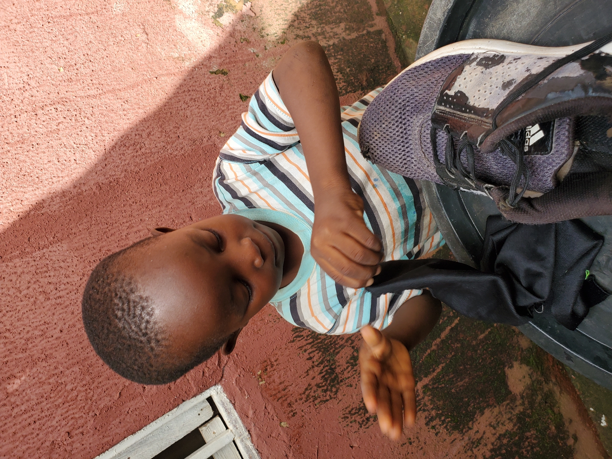 A young Black child, with short, dark hair and brown eyes, is shown from the shoulders up, focused downwards. The child wears a horizontally striped t-shirt in shades of blue, white, orange, and black. Their hands are engaged with a black piece of fabric. In the foreground, to the right, rests a partially visible dark grey athletic shoe with a white sole and black laces. The child appears to be outdoors, as indicated by the textured, reddish-brown wall behind them, which shows signs of weathering and some green mossy patches. A sliver of a white window frame is visible to the left. The lighting suggests it is daytime, with a distinct shadow cast across the wall and the child from the upper left. The scene is likely in Jos, Nigeria, given the architecture and typical outdoor environment.